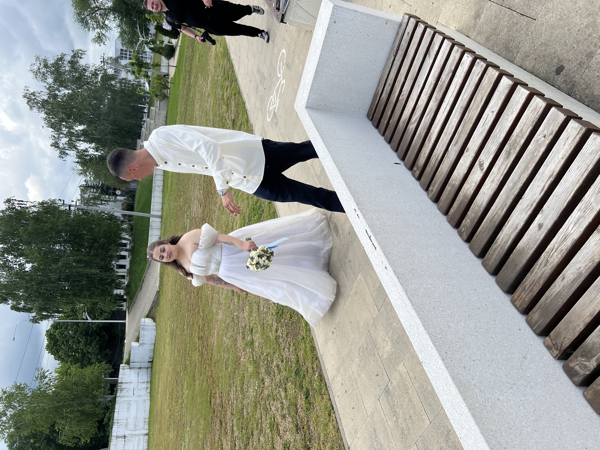 The image shows a wedding photo in Goryachiy Klyuch, Russia. The bride, wearing a white dress and holding a bouquet, stands next to the groom, who is in a white shirt and dark pants. They are positioned beside a modern bench with a concrete base and wooden slats. The background includes a green lawn, trees, and buildings, suggesting an urban park setting. Other people are present in the background. The sky is overcast. The photo appears to be taken at an angle.