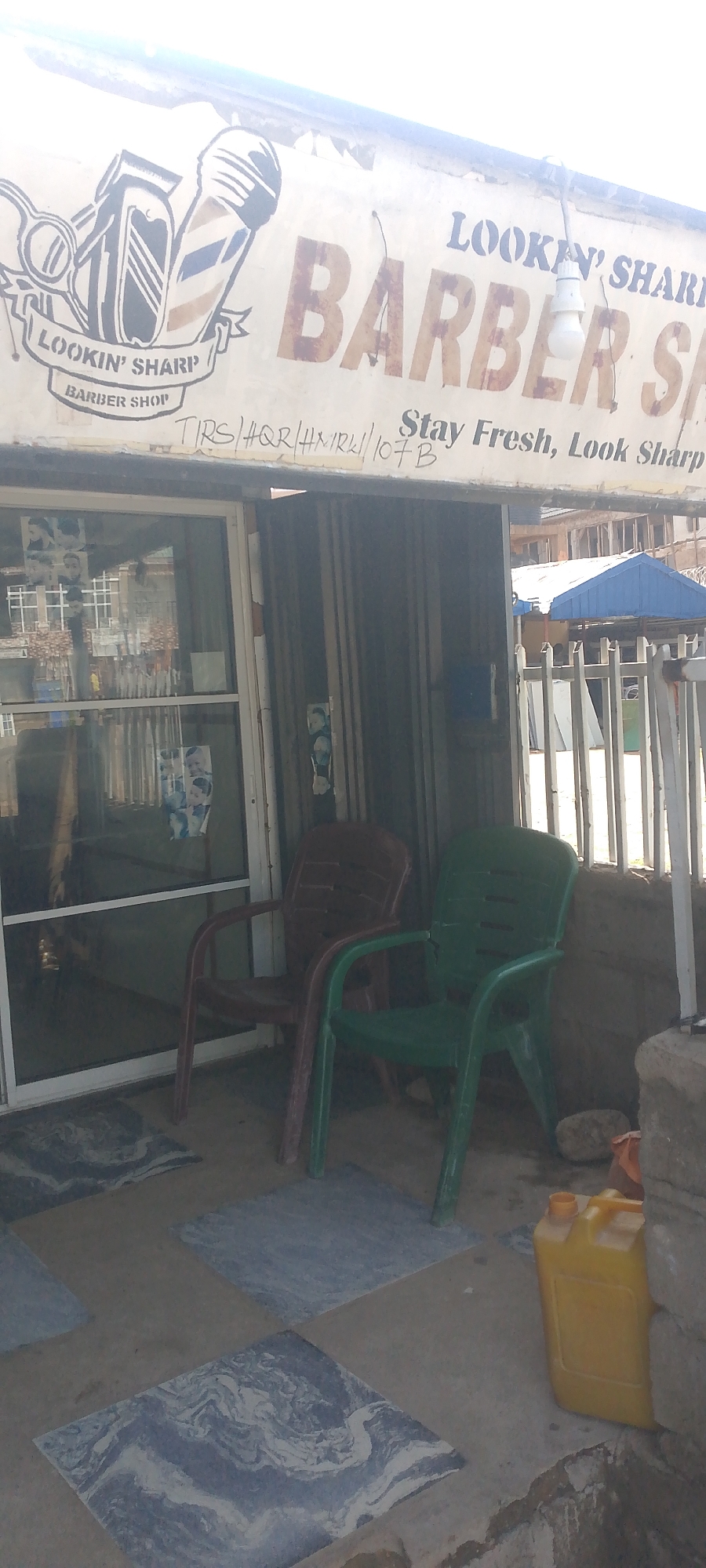 This is a medium-shot, eye-level outdoor photograph of a barber shop in Jalingo, Nigeria. The barber shop's sign, which reads "LOOKIN' SHARP BARBER SHOP," is prominently displayed above the entrance. The sign features a graphic of a barber's clipper and scissors. Below the main sign, there is handwritten text, which appears to be a name or code. To the right of the sign, the slogan "Stay Fresh, Look Sharp" is also visible.
The shop's entrance consists of a glass door and window, through which several framed pictures of hairstyles are visible. Outside the entrance, two plastic chairs, one brown and one green, are placed on the ground. The ground is partially covered with square tiles featuring a marbled pattern. To the right of the chairs, a yellow plastic jerrycan is situated next to a brick wall. The background shows a glimpse of buildings and a blue canopy, suggesting an urban or semi-urban environment. The lighting suggests it is daytime, with bright sunlight creating some glare. There are no people clearly visible in the immediate foreground or engaging in any actions. The overall impression is of a functional, small business establishment.