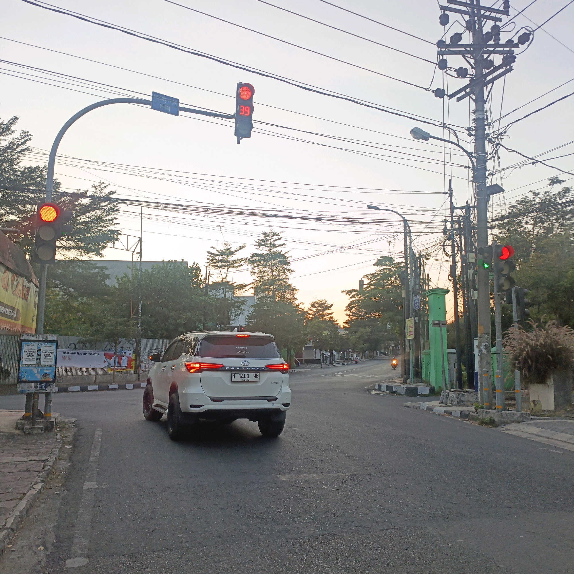 The image depicts a street scene in Salatiga, Indonesia, likely during sunset given the warm, diffused light. In the foreground, a white Toyota Fortuner SUV is driving away from the camera on a paved road. The vehicle's brake lights are illuminated, suggesting it may be slowing down or stopping. To the left of the road is a sidewalk with some advertisements and a traffic light displaying red, with a countdown timer showing "39." Further down the street, another traffic light is visible, also showing red and green lights. Numerous utility poles with a complex web of wires are prominent features throughout the scene. Trees and buildings line the sides of the road, some with lush green foliage. The sky is a pale orange and yellow, indicating the sun is low on the horizon. The overall impression is a typical urban street at dusk, with traffic and infrastructure elements typical of Indonesia.