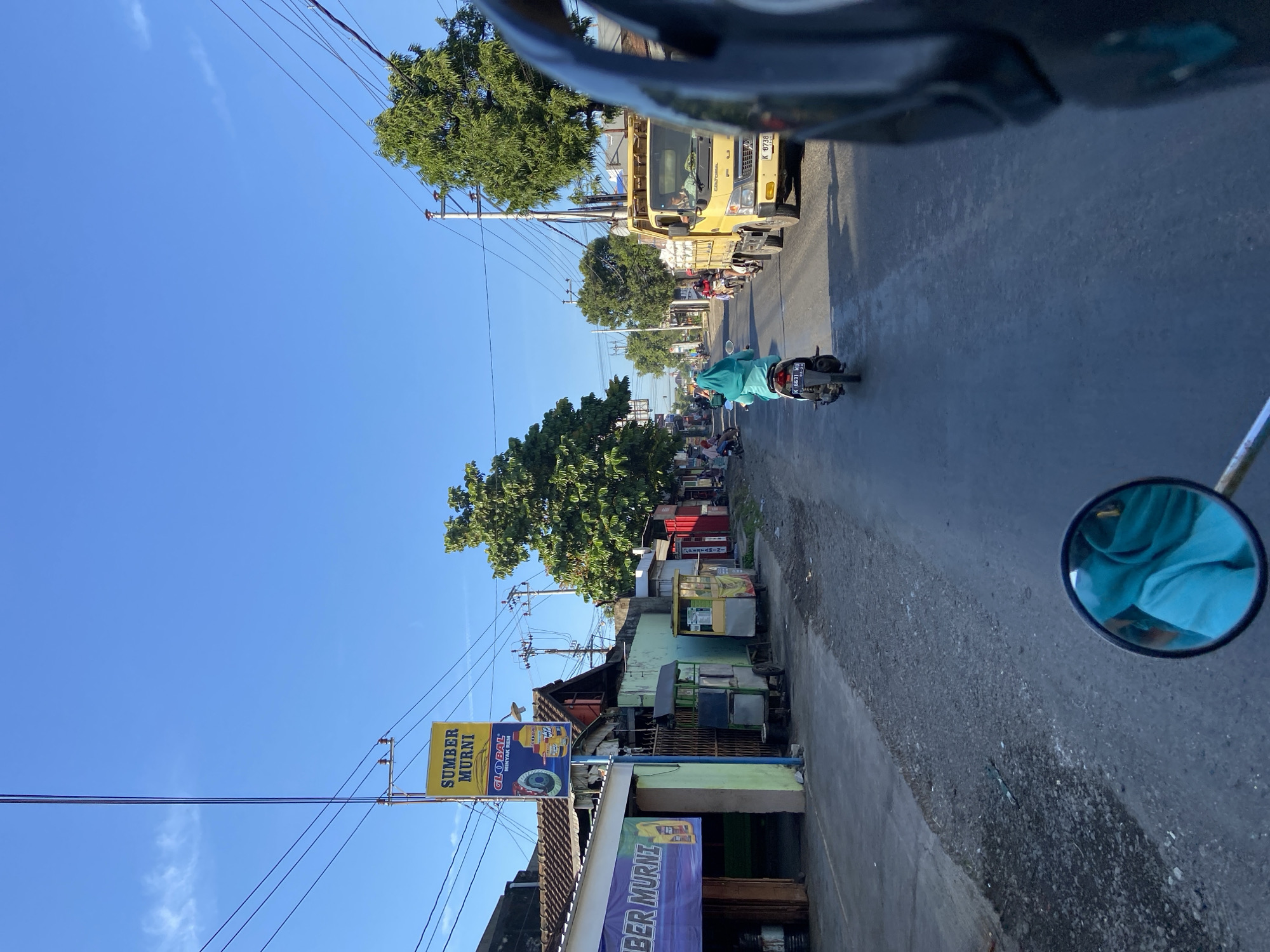 This is a street-level, first-person perspective photograph taken in the city of Margoyoso, Indonesia. The scene depicts a street with businesses lining the left side, including a storefront with a purple banner reading "SUMBER MURNI." A yellow sign above it also reads "SUMBER" and "MUSNI." Further down the street, other small businesses and street vendors are visible.
The road itself is paved and appears to be moderately trafficked. In the foreground, a motorcycle's rearview mirror is prominently visible, reflecting a person wearing a blue head covering. Ahead on the road, a person on a scooter, also wearing a blue head covering, is riding away from the camera. Further up the road, a yellow truck is traveling in the same direction.
The sky is clear and blue, indicating a sunny day. Trees line the right side of the street, and utility poles with numerous wires are scattered throughout the scene. The overall impression is that of a typical, busy street in a small Indonesian town.