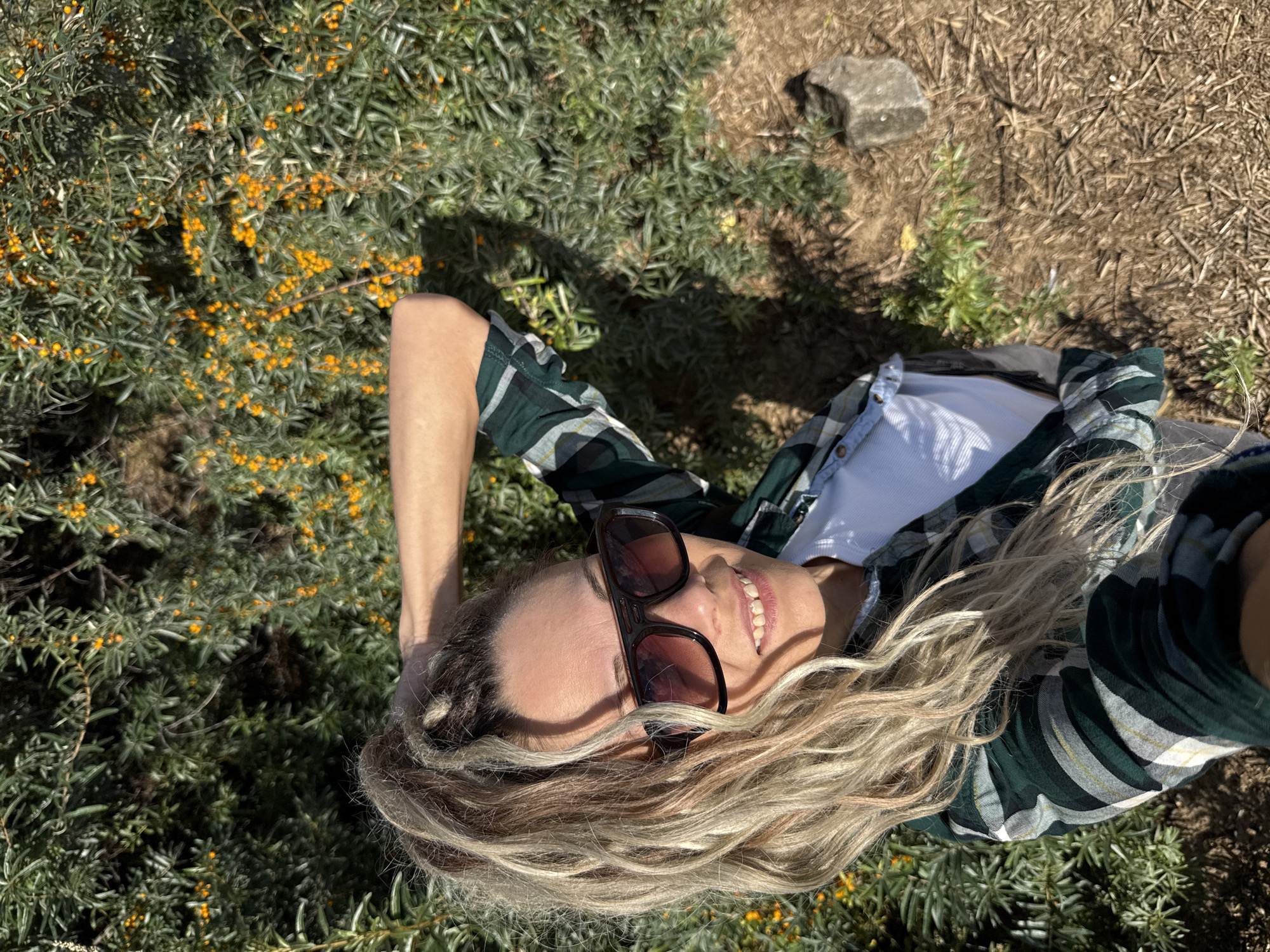 In the city of Rusanovo, Russia, a woman is captured in a selfie outdoors, positioned against a backdrop of lush green foliage interspersed with numerous small, bright orange berries, likely sea buckthorn. She is wearing a green and white plaid button-down shirt over a white t-shirt and dark-rimmed sunglasses with tinted lenses. Her blonde hair is styled in a high ponytail, and she has one arm raised, with her hand resting on the back of her head. The lighting suggests it is daytime, with distinct shadows cast on the ground. The ground itself is covered in dry grass and twigs, with a small, grey rock visible to the right of the frame.