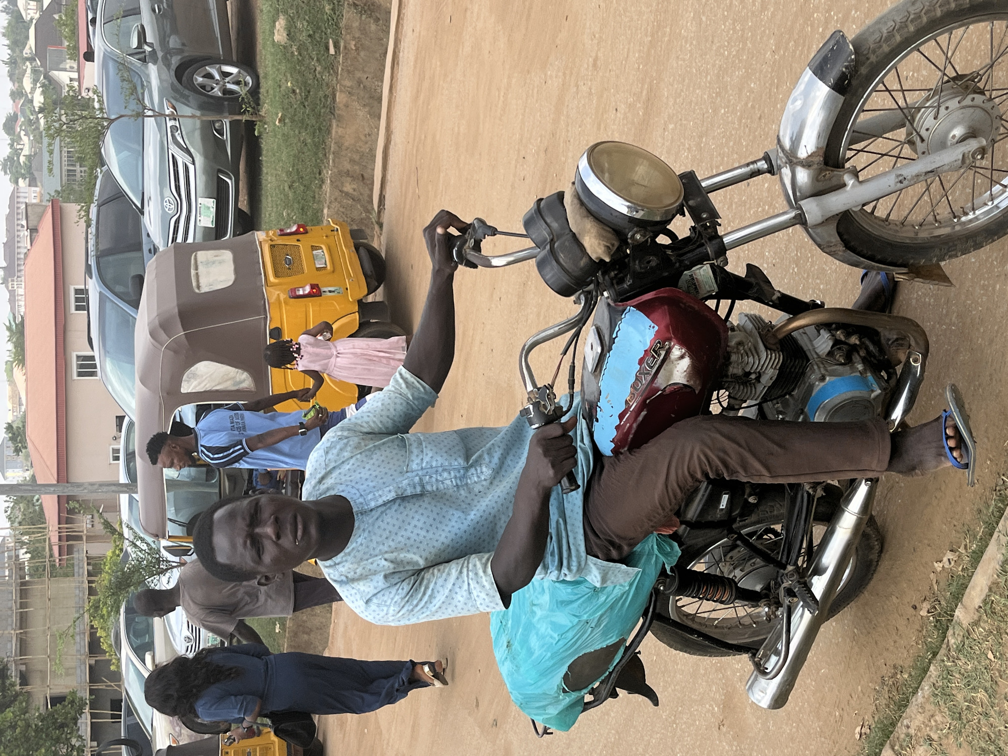 A man is sitting on a motorcycle in Abuja, Nigeria. The motorcycle is old and has a red gas tank with blue accents. The man is wearing a light blue patterned shirt and brown pants. He is holding the handlebars of the motorcycle and looking at the camera. There is a blue plastic bag hanging on the back of the motorcycle. Behind the man, there is a yellow auto rickshaw and several cars parked along the side of the road. There are also several people walking in the background. The background shows buildings.