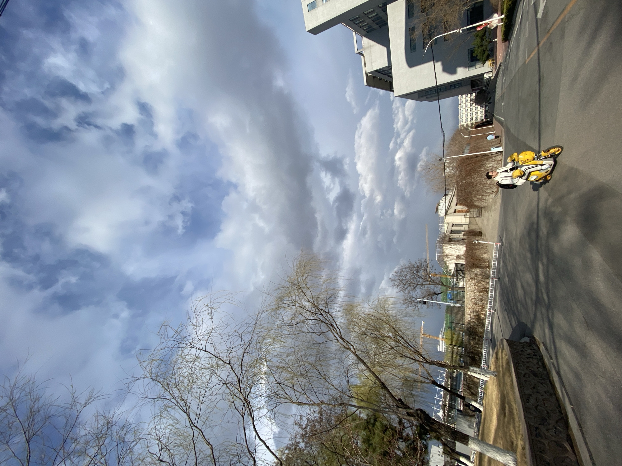 A low-angle view shows a sunny street scene in Zhongtun, China. A man is riding a yellow scooter on the right side of the road. Beside the road is a curb and trees. In the background is a partially seen crane, buildings, and light poles. The sky is mostly blue with streaks of clouds.