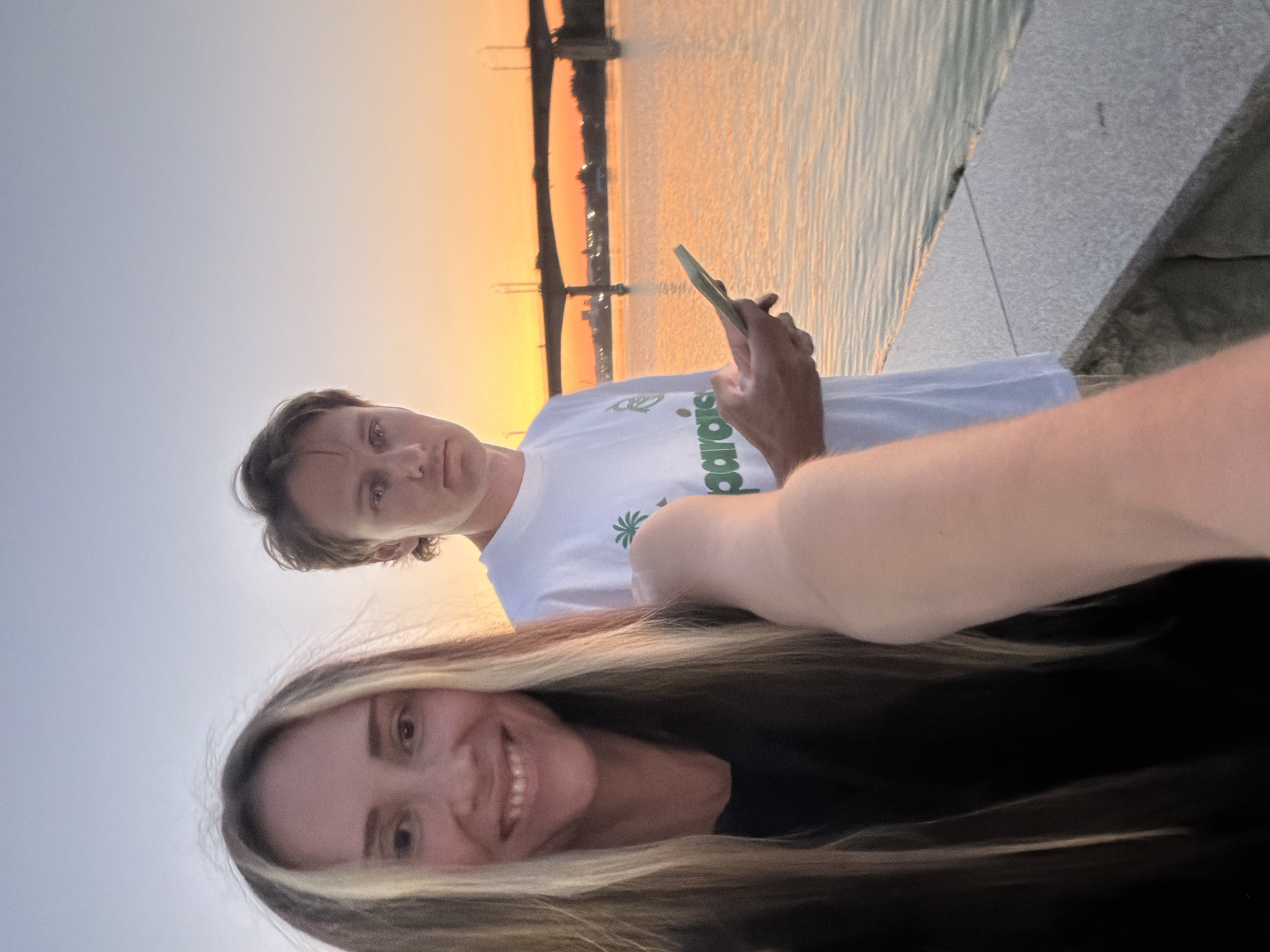 This image shows two individuals, a woman and a man, posing for a selfie by a body of water during sunset. The woman is in the foreground, smiling and looking at the camera. Her blonde hair is long and partially obscures her face. She is wearing a black top. The man is standing behind her, looking towards the camera with a neutral expression. He is wearing a white t-shirt with green graphics. He is holding a green smartphone in his right hand.
They are standing on a stone embankment. In the background, a bridge spans a wide river, and the sky is illuminated with the warm colors of a sunset. The scene appears to be taking place along the Volga River embankment in Kamyshin, Russia, given the context provided by the prompt. The water reflects the orange and yellow hues of the sky.