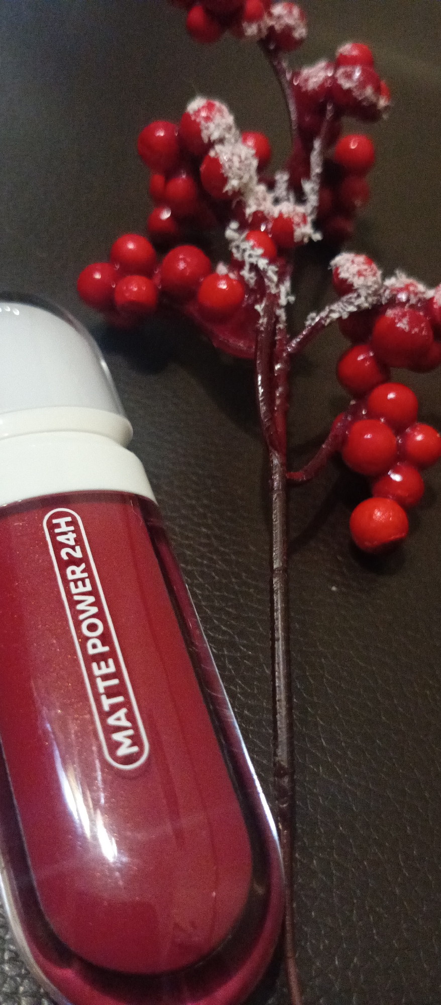 A close-up, angled, indoor shot shows a container of red liquid cosmetic product (likely lip gloss or lipstick) next to a red berry sprig against a dark leather-like surface. The container has a clear, elongated rectangular base with a white top. A white label with red lettering "MATTE POWER 24H" is visible on the container. To the right of the container is a sprig of red berries, some of which are dusted with white, possibly artificial, snow-like material. The berries are arranged in clusters along the dark red sprig. The surface appears to be a dark brown, possibly black, textured material, likely leather or faux leather.