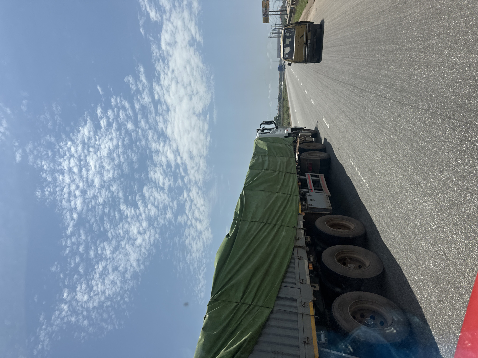 In Magboro, Nigeria, a multi-axle truck covered with a green tarp is traveling on an asphalt highway. The truck, positioned in the left foreground, has its rear axles and wheels visible. Further down the road, a yellow minivan occupies the right lane, with several individuals visible inside. The highway extends into the distance, bordered by a grassy median and vegetation on the right. The sky above is a clear blue, dotted with scattered white clouds. The overall scene depicts highway traffic under a bright sky.