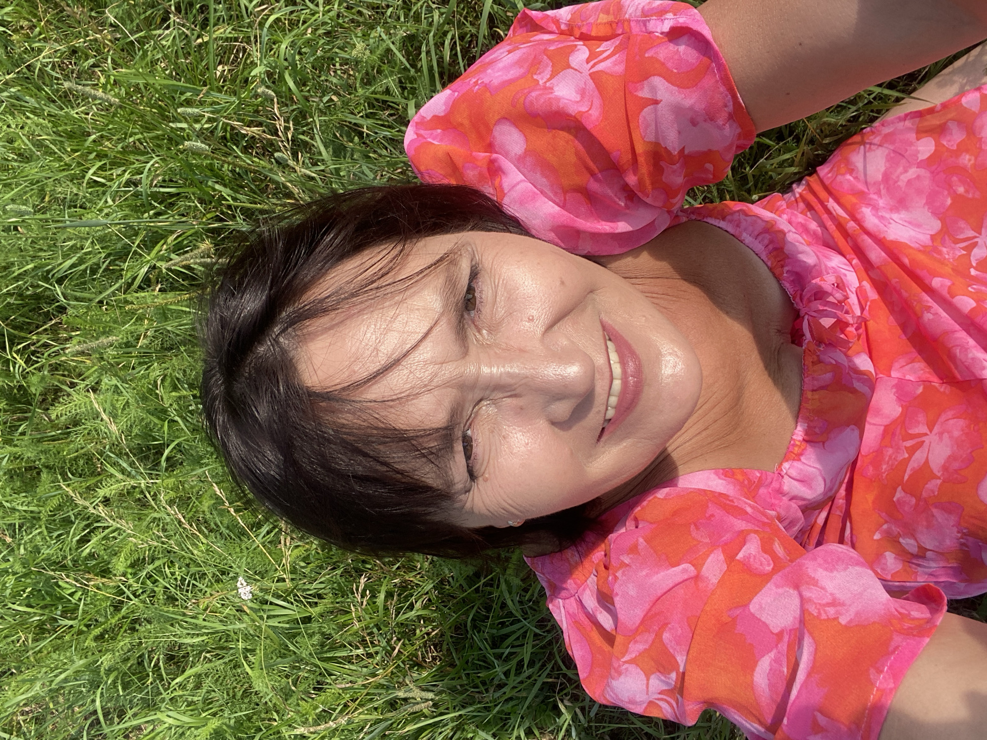 A woman is lying on grass in a field, looking up at the camera. She has dark hair and is wearing a pink and orange floral dress. The background is out of focus green grass. The image appears to have been taken on a sunny day. There is no information to suggest this image was taken in Novokuznetsk, Russia.