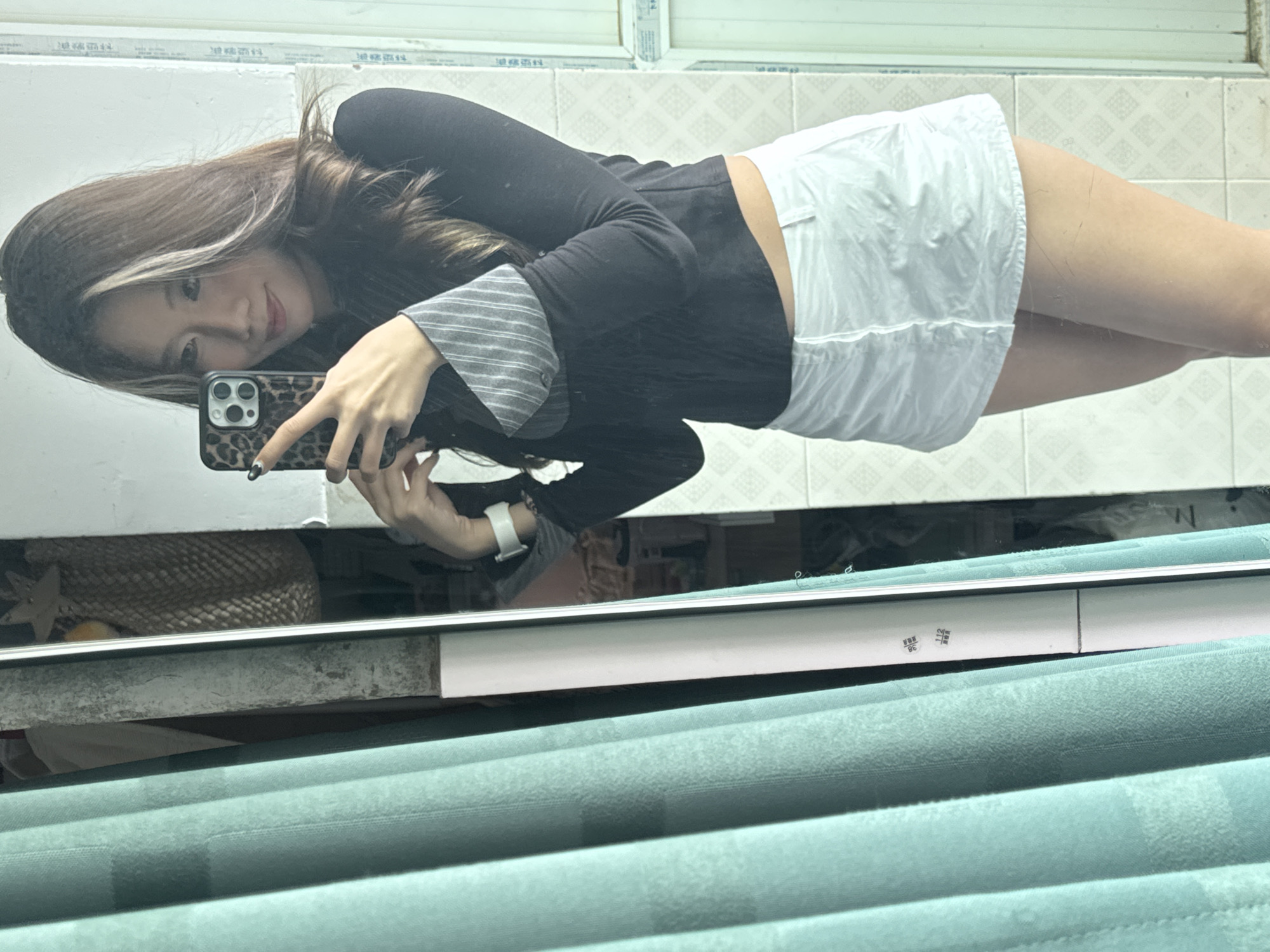 A young woman takes a mirror selfie. She holds a phone with a leopard print case and wears a black top with striped sleeves and a white skirt. The background consists of a tiled wall. The foreground shows a green blind and part of the room where the selfie is taken. The image was likely captured indoors, and the location is possibly Nan'an District, China, though this cannot be definitively determined from the image alone.