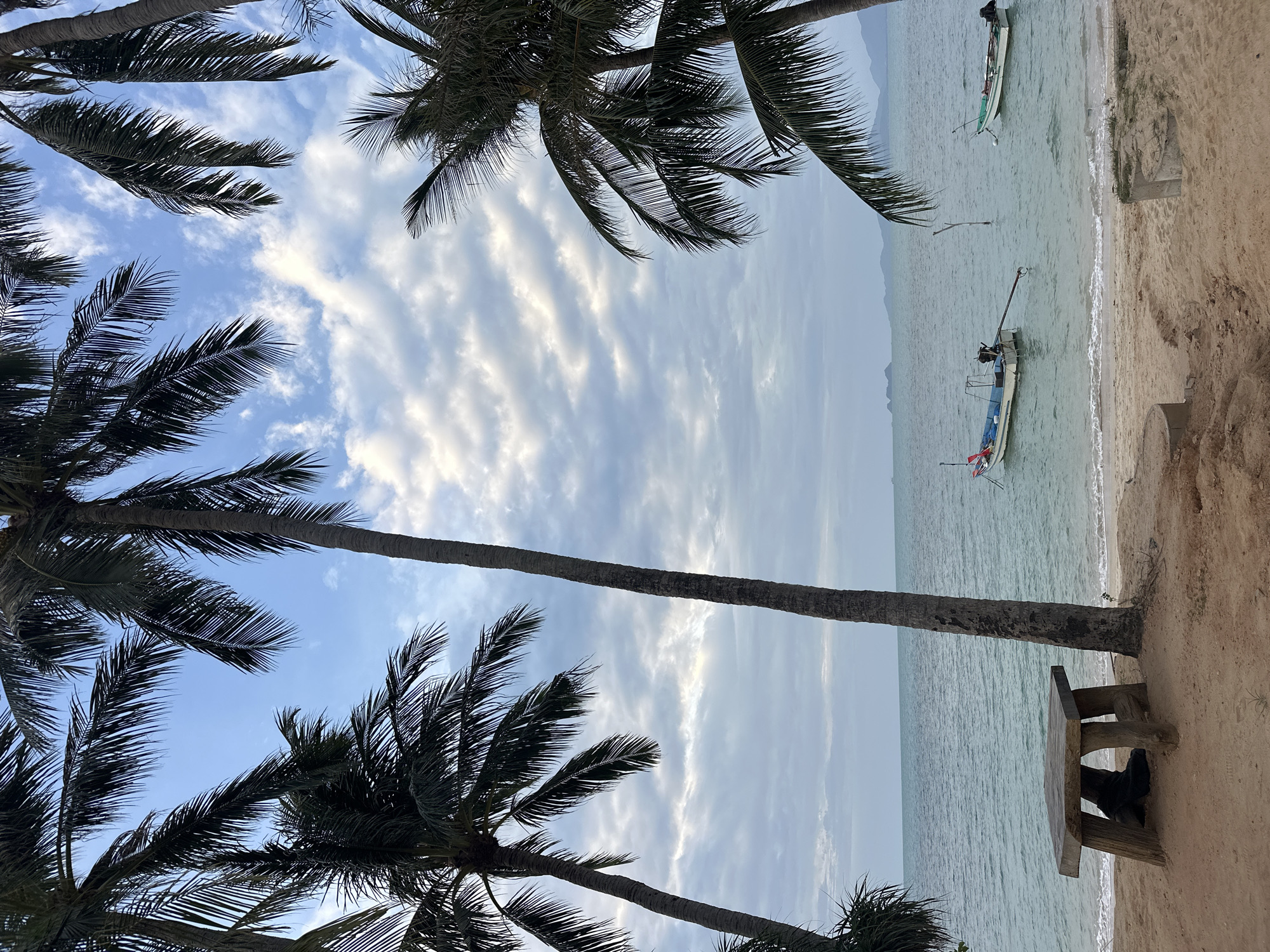 A serene beach scene unfolds in Ko Samui District, Thailand, framed by the silhouettes of tall palm trees. The foreground features a rustic, wooden table with two plank seats, positioned on the sandy shore. The turquoise water of the sea gently laps at the beach, and two small fishing boats are anchored offshore. In the distance, a hazy coastline with rolling hills is visible. The sky above is a soft blue, adorned with scattered white clouds that create a sense of depth and tranquility. The overall atmosphere is peaceful and idyllic, indicative of a tropical paradise.