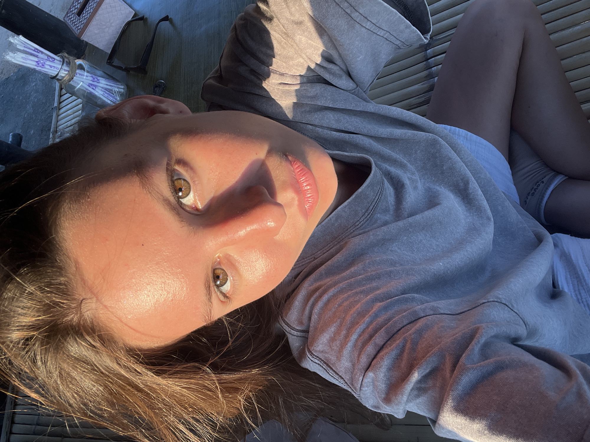 A close-up, high-angle selfie shows a young female with brown hair and brown eyes. She is wearing a grey t-shirt and white shorts. The scene appears to be outdoors, possibly on a wooden platform. In the background, there are some straw-like materials, a glass bottle with purple straws, sunglasses, and a tissue box. The lighting suggests it is either sunrise or sunset, casting a warm glow on her face.