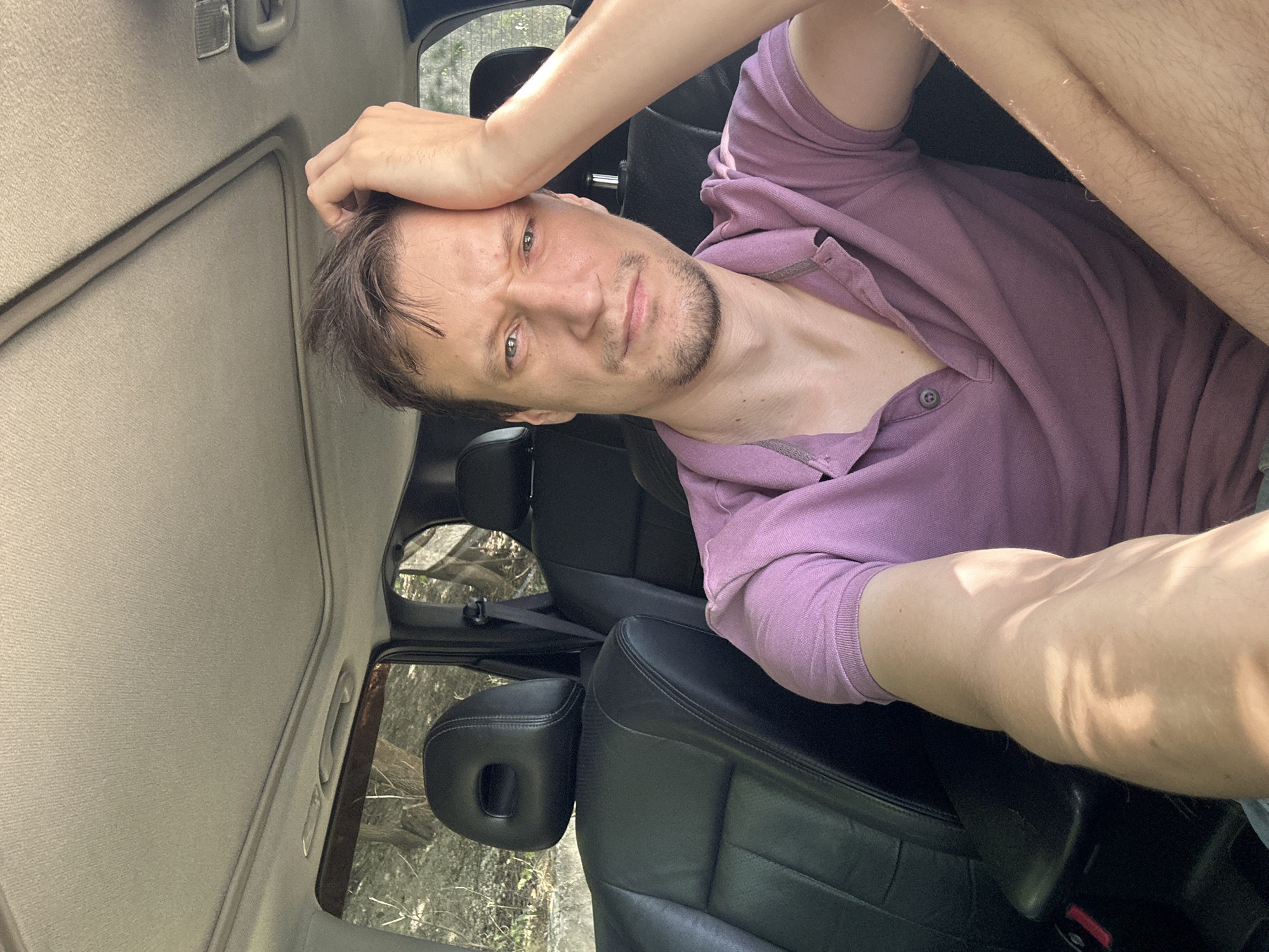 A man is sitting in the driver's seat of a car, taking a selfie. He has light brown hair and a light beard. He is wearing a purple polo shirt. His right arm is bent, and his hand is touching his forehead. The car appears to be parked outdoors, with trees and foliage visible through the windows. The interior of the car has black leather seats and a sunroof. The image was taken during the daytime. There is no indication of the location being in Makiv, Ukraine, or any specific city.