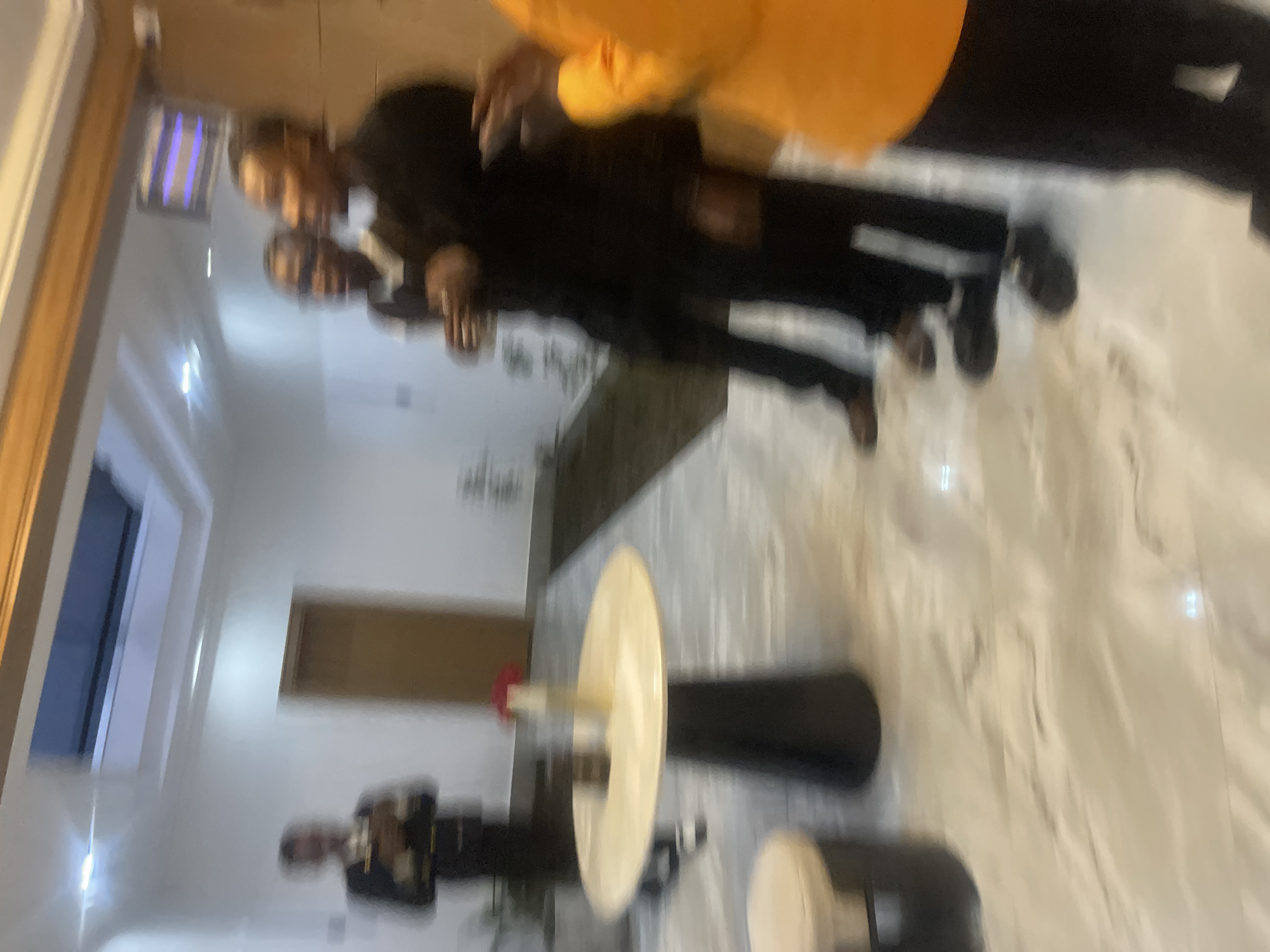 The image depicts an indoor scene, possibly a reception area or hallway, with a marble-tiled floor. Several individuals are present, all appearing to be men. In the foreground, a person in an orange garment is partially visible on the right. To their left, a person in a black suit stands. Further back, to the left of the suit-clad figure, two other men in dark attire are standing. On the far left, a man in what appears to be a uniform or formal attire, possibly holding an object, is visible near a door. A round table with a light-colored top and a dark pedestal base is situated in the middle ground, with a vase containing a red flower and another object on it. The lighting suggests an interior space, with a skylight visible in the ceiling. The overall impression is one of people gathered in a contemporary indoor setting. There is no specific visual information that places this scene within Gura Top, Nigeria.