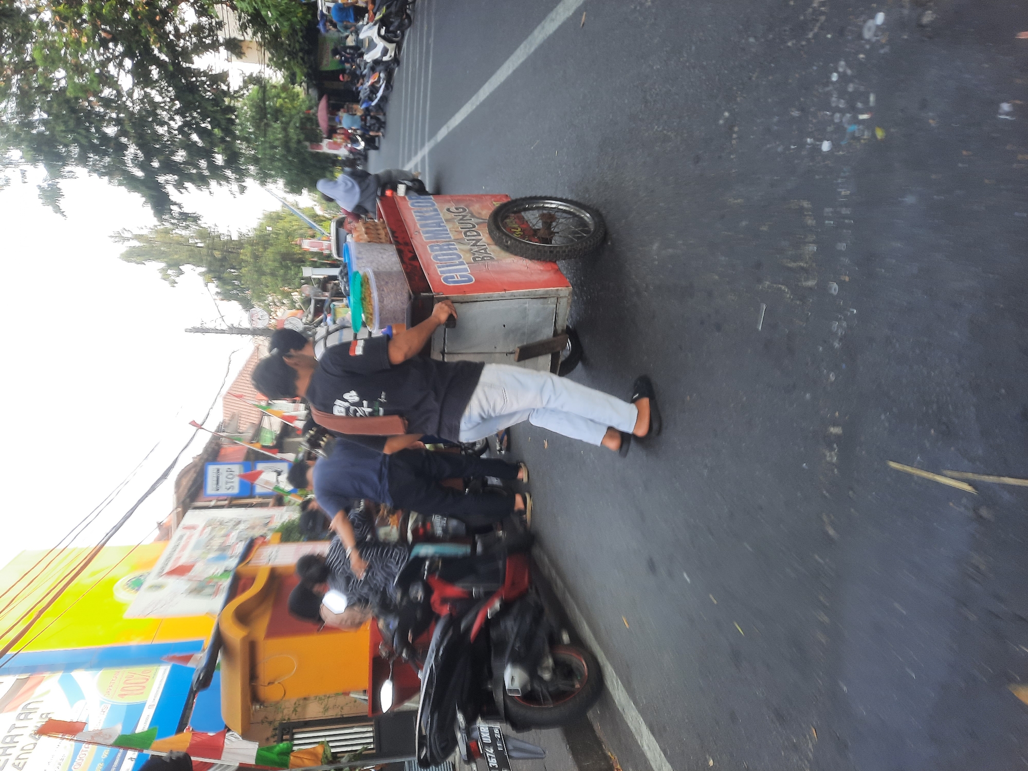 An outdoor street scene in North Jakarta, Indonesia, depicts a busy intersection with several people and vehicles. A man wearing a black t-shirt and light-colored pants is pushing a food cart with red and silver sides, which displays text that appears to read "CIUOR MAKLO" and "BANDUNG". The cart is filled with containers of food items. To the left of the cart, a red and black motorcycle is parked, with a license plate visible that reads "3674 UXR". In the background, other individuals are seen on motorcycles and on foot. A "STOP" sign is mounted on a pole. The environment includes buildings with colorful signage and trees lining the street. The overall atmosphere is dynamic and suggests daily commerce.