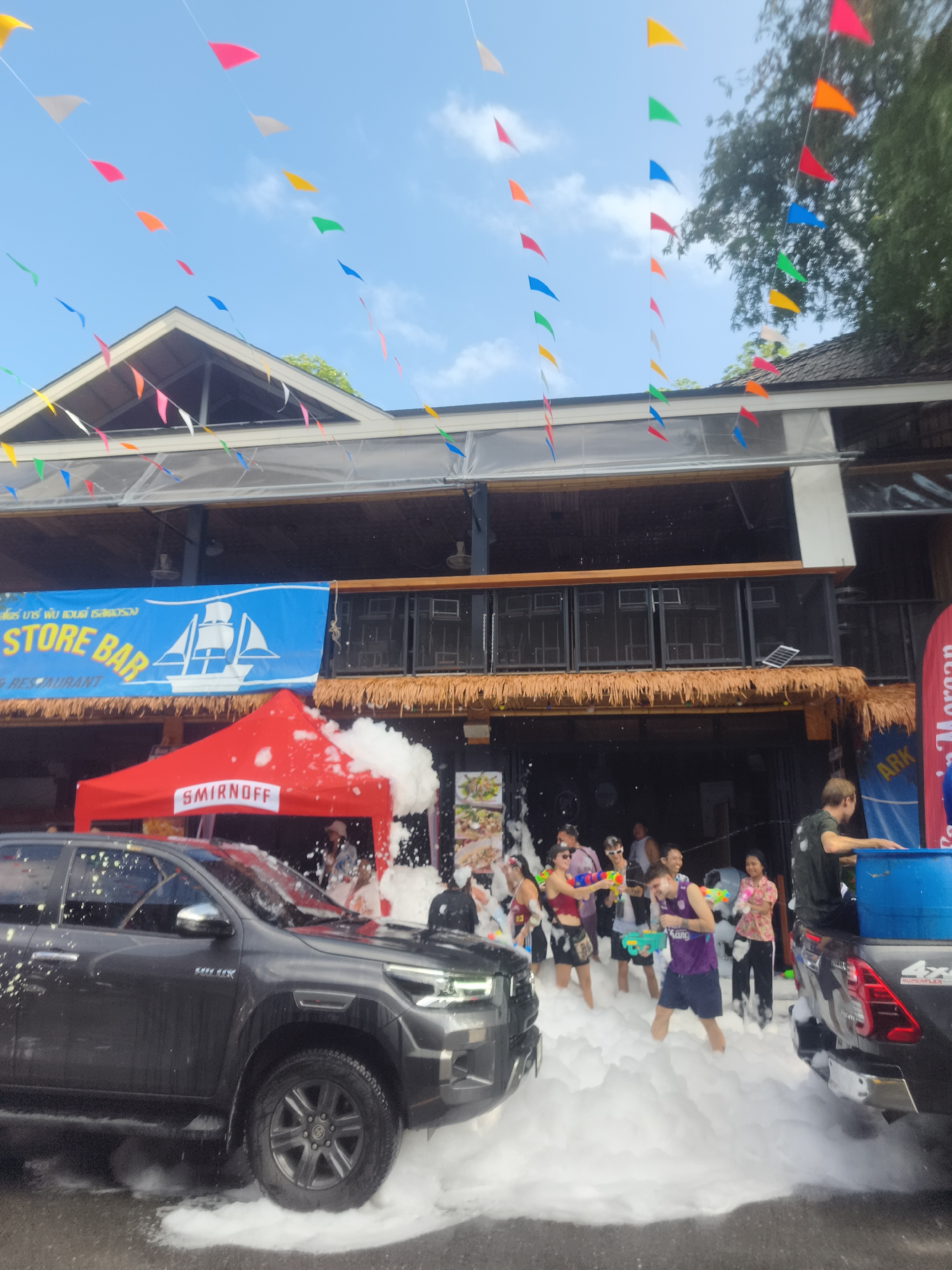 The image captures a lively street scene in บ้านบางรักษ์, Thailand, likely during a festival or celebration. A two-story building serves as the backdrop, adorned with colorful pennants strung overhead. A banner reading "STORE BAR & RESTAURANT" is visible above the ground floor, which features a thatched roof overhang. A red Smirnoff tent is set up near the building's entrance.
Numerous people are engaged in water-related activities, using water guns and buckets, amidst a thick layer of white foam covering the street. Two dark-colored pickup trucks are parked on either side, partially obscured by the foam. One truck is branded with "HILUX" and the other with "4x4 SUPERFLEX" and "ARK Morgan".
The overall atmosphere suggests a festive and playful environment.