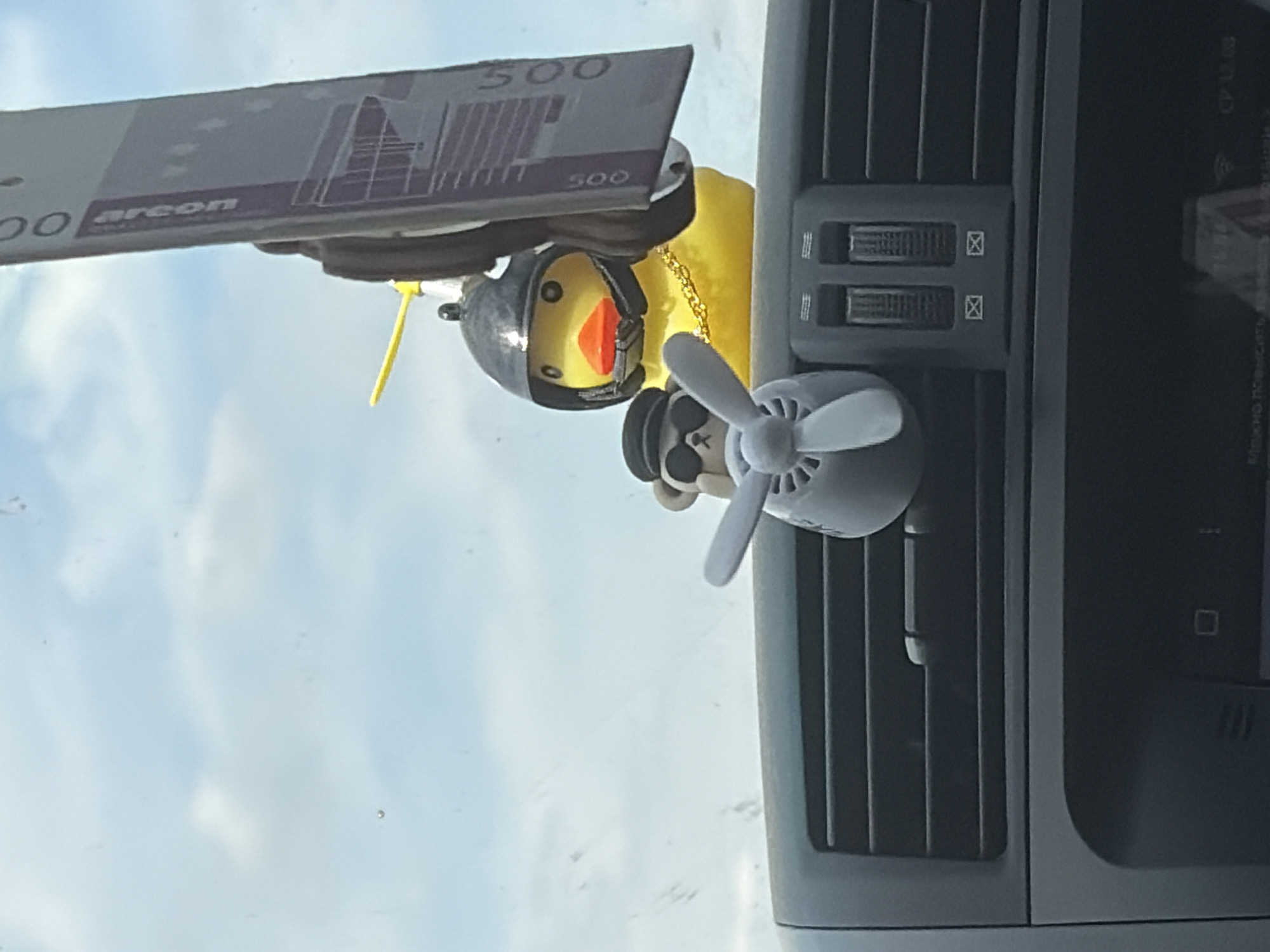 The image depicts a yellow rubber duck wearing a black helmet and a gold chain, positioned on a car's dashboard. To the left of the duck, a small grey bear wearing sunglasses and a cap is visible. In front of these figures, a small grey airplane propeller ornament is attached to the air vent. The background shows a cloudy sky. The scene is consistent with a vehicle interior in Kruglovka, Russia, though the specific location within the city is not discernible.