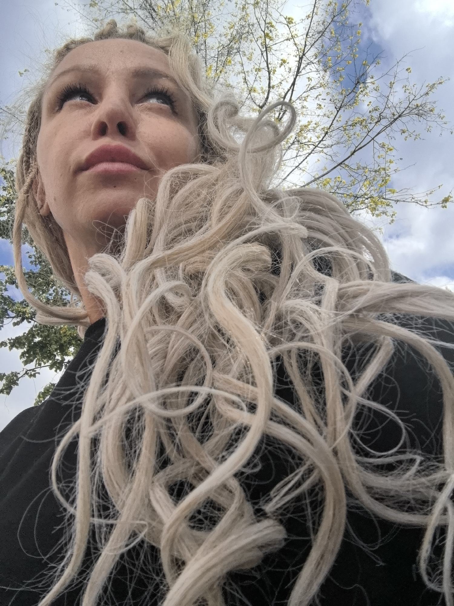 This is a low-angle, close-up, portrait-style photograph of a woman with blonde hair styled in dreadlocks. She is wearing a black garment. Her face is looking upwards, and her eyes are open, directed towards the sky. The background consists of tree branches with small yellow leaves against a cloudy blue sky. The image does not contain any visual information that would allow for the identification of a specific location within the city of Chaikovsky, Russia, or any other city.