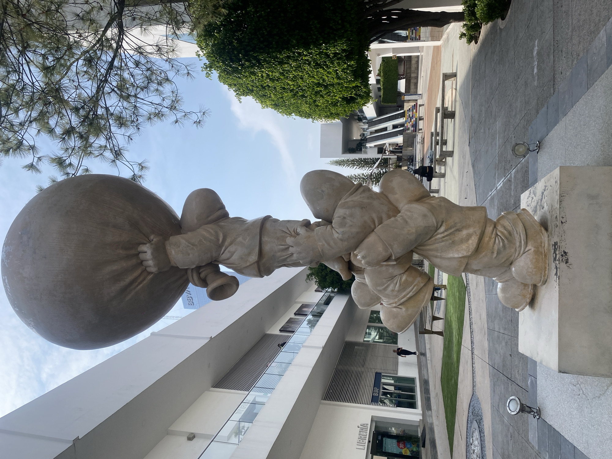 The image displays a large, stylized sculpture of three figures situated in a modern urban plaza under a clear blue sky. The sculpture, crafted from a light-colored, smooth material resembling stone or concrete, features rounded, almost childlike forms. The leftmost figure is prominently shown carrying a massive, spherical sack over its shoulder, gripping it with both hands. Two other figures are positioned to its right, appearing to be linked or assisting in the collective effort of transporting the heavy load.
The setting is a contemporary public area with minimalist architecture. To the left and behind the sculpture stands a multi-story white building with extensive glass windows and horizontal louvers. At ground level, a storefront is visible with the text "LIBRERIA" (Bookstore) and a "30% OFF SALE" sign. Nearby, the acronym "BUAP" is present on another glass panel. In the distance, a building bearing the "BBVA" logo can be faintly seen.
The plaza itself features varied patterns of grey and beige paving stones, interspersed with sections of green grass and several modern benches. Large, lush trees, including some with pine-like branches, frame the upper parts of the scene against the bright, clear sky. The ample natural light indicates it is daytime, likely morning or afternoon, under pleasant weather conditions. A few individuals are visible in the background, walking or standing, contributing to the sense of a functional, public space. The sculpture evokes themes of cooperation or shared responsibility within this clean, modern environment.