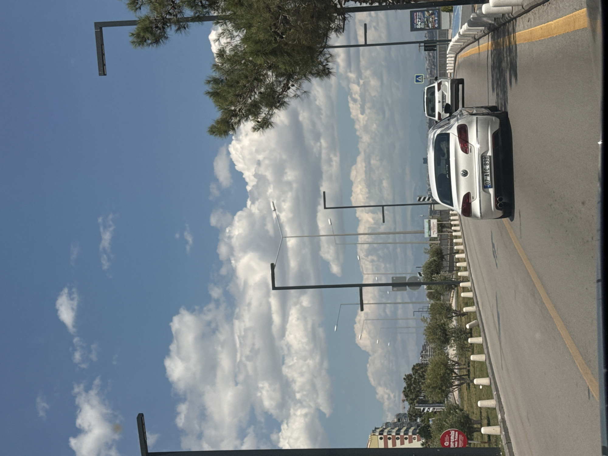 The image, likely taken from a vehicle, shows a road with a yellow dividing line in Konyaaltı, Turkey. A white car with the license plate "07 ATC 898" is directly in front, followed by another white car. Streetlights line the road, which is bordered by a grassy area and small trees. The sky is partly cloudy. Buildings are visible in the distance.