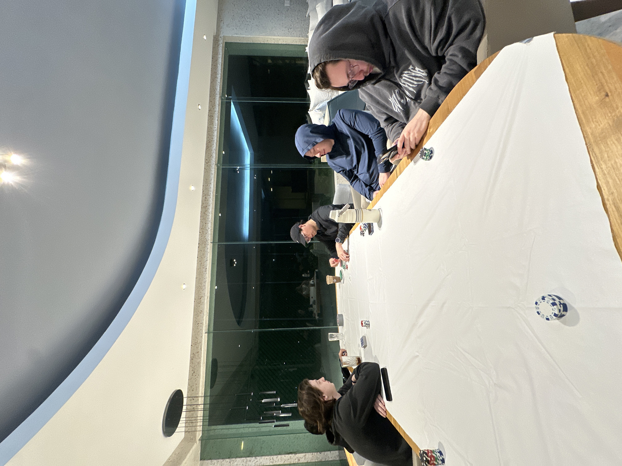 Four individuals are sitting around a large, oblong table covered with a white cloth, with scattered poker chips and glasses. The table has a light wood edge. The individuals appear to be engaged in a poker game. Three are wearing hoodies. One is wearing a baseball cap. Two of the four individuals are holding cell phones. The scene takes place indoors, next to a large window with a green outdoor area visible. The ceiling is domed. The location is Naucalpan de Juárez, Mexico.