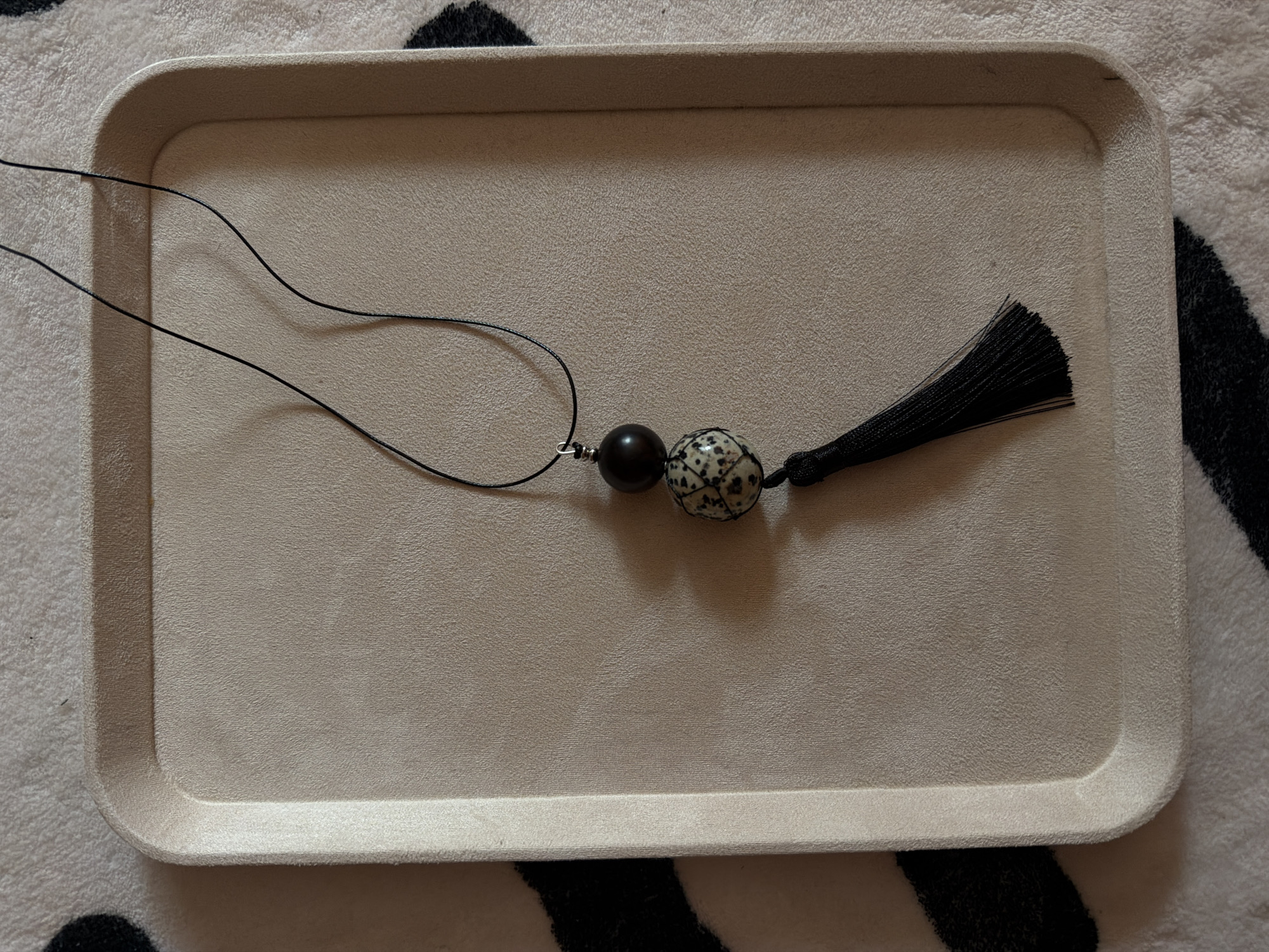 A flat lay image shows a necklace with a black cord, a dark brown bead, and a stone bead with a black and white speckled pattern, hanging from a black tassel. The necklace rests on a beige tray with rounded corners. The tray is placed on a textured rug with a black and white abstract pattern. The image is taken from a top-down perspective. The specific location in the city of Khimki, Russia, cannot be determined from the image.