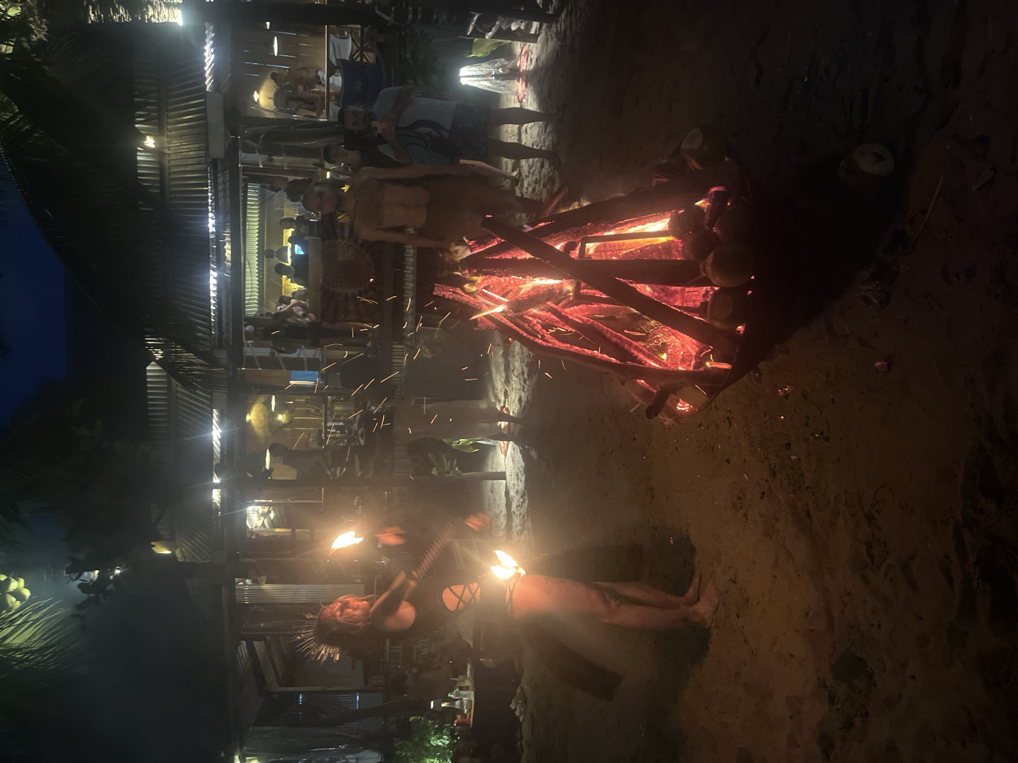 An outdoor nighttime scene on a sandy beach, likely in Đà Nẵng, Vietnam, features a fire performance. In the foreground, a woman in a dark, cutout swimsuit and a headdress is mid-performance, holding flaming torches in both hands. Sparks from the fire are visible in the air. A large bonfire is in a metal basin to the right, containing logs and coconuts. Behind the performers, a beachside establishment with a corrugated metal roof is lit up, with several people visible inside and standing around. The lighting is dim, with the primary illumination coming from the bonfire and torches. Palm trees are visible in the dark background. The sand is the dominant ground cover.