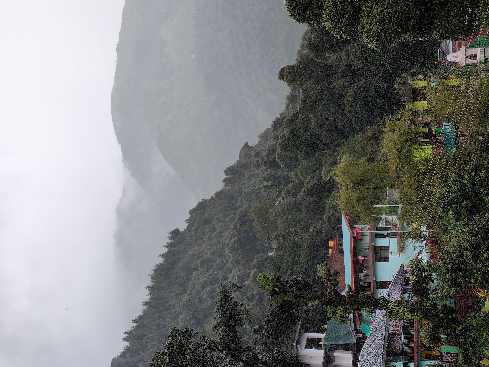 This is a vertical, eye-level, medium shot of a hillside village in Haini, India, on a cloudy and overcast day. The dominant colors are shades of green from the dense foliage and teal from the buildings. In the foreground, several multi-story buildings are nestled into the slope of a lush, tree-covered mountain. The architecture features a mix of traditional and modern elements, with balconies, tiled roofs, and brightly colored facades, including teal, pink, and yellow. Clothes are seen drying on a clothesline on one of the balconies. Further up the mountain, the trees become denser, and the mist begins to obscure the higher peaks. The overall atmosphere is serene and somewhat mysterious, with the mist lending a dreamlike quality to the landscape. The image appears to be taken during monsoon season, as indicated by the heavy cloud cover and possible rain streaks visible.