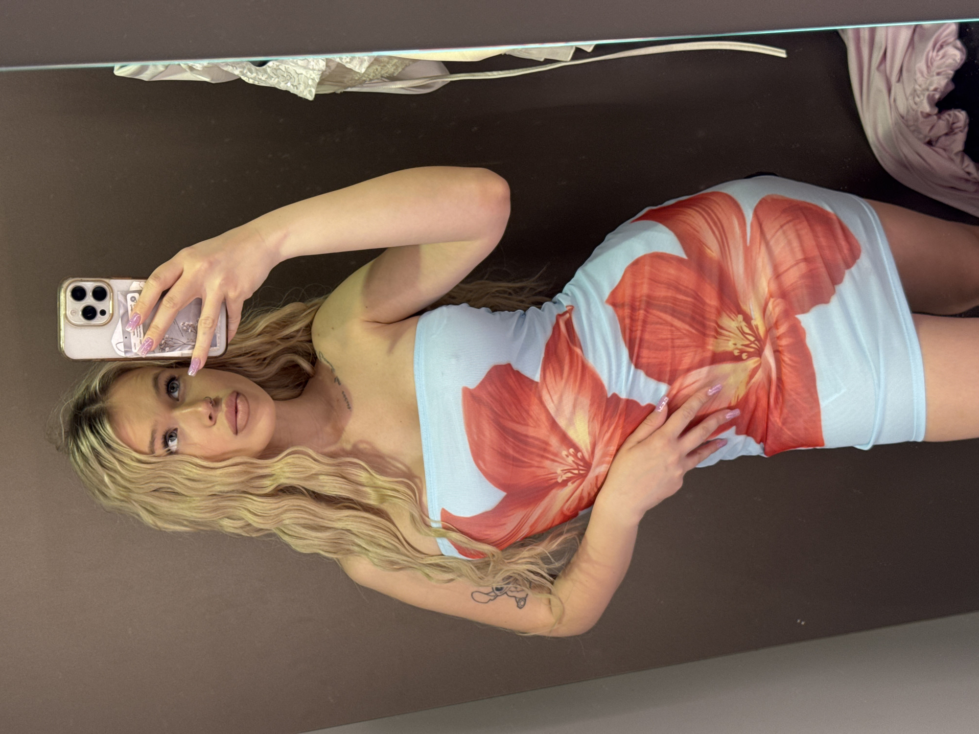 A young woman with long, wavy blonde hair and light-colored eyes is taking a mirror selfie in what appears to be a fitting room. She is wearing a light blue, strapless mini-dress with a large orange and red floral print. Her left arm is bent, and her hand is resting on her hip, while her right hand holds up her phone to take the picture. The background is a plain brown wall, and a mirror reflects the woman and the room. A glimpse of clothing is visible to the right of the mirror, suggesting the presence of a rack or display. The overall impression is a casual, personal moment captured in a retail or changing space. The image does not contain any specific information or visual cues to indicate a location in Kudrovo, Russia.
