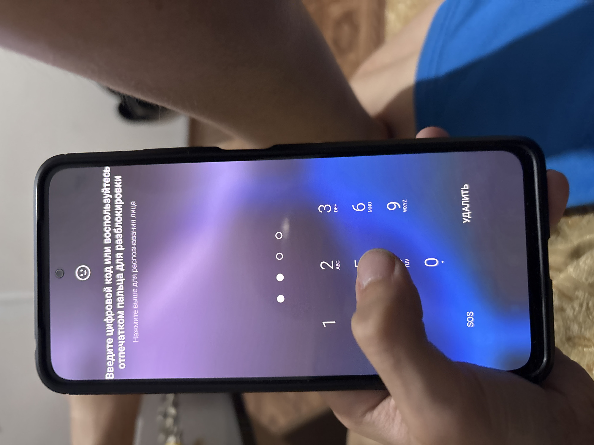 The image shows a close-up of a smartphone screen displaying a Russian language lock screen. The text on the screen reads "Enter a numeric code or use fingerprint for unlocking" and "Tap above for face recognition." Below this, there are four white dots, likely indicating a password or pattern entry progress. A numeric keypad is visible with numbers 1 through 9, and a 0 button. A finger is pressing the number 5 key. The bottom of the screen shows "SOS" on the left and "DELETE" on the right. The phone is held by a person, whose hand is partially visible. The background is blurred and appears to be indoors, with a surface that resembles a textured fabric, possibly a bed or sofa. There is no visible indication of the city of Grozny, Russia in the image.