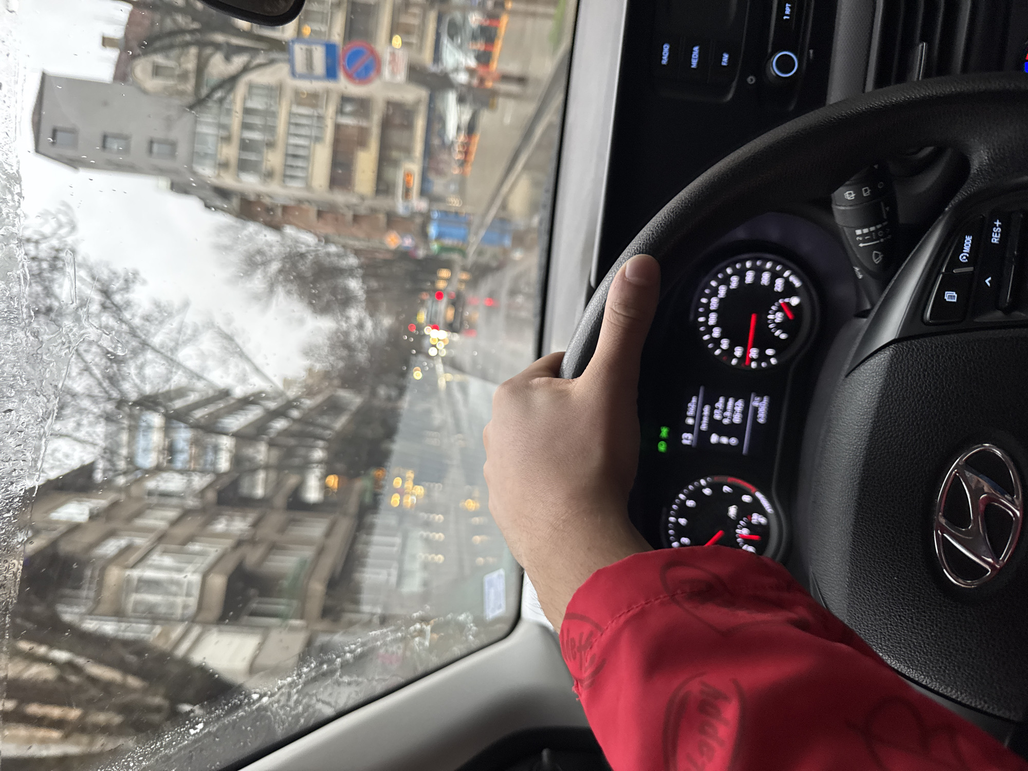 The image depicts the interior of a Hyundai car, with a driver's hand visible on the steering wheel. The Hyundai logo is visible on the steering wheel. The dashboard displays various information, including the speedometer, RPM, and other vehicle data. The windshield is wet, indicating rain or snow. The exterior view through the windshield shows a street scene with buildings and traffic. A traffic sign can be seen in the distance. The buildings appear to be located in the city of Sofia, Bulgaria, based on the architectural style and traffic signs. A red jacket sleeve is visible in the bottom right corner.