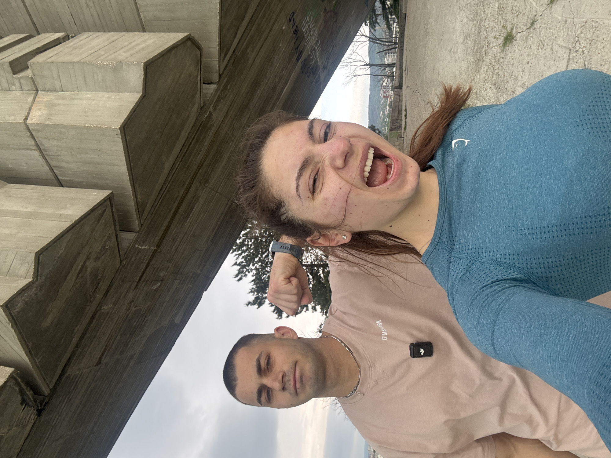 Two individuals, a man and a woman, are positioned under a concrete overpass or bridge structure. The location is likely Varna, Bulgaria due to architectural style of concrete. The woman, in the foreground and to the right, is wearing a blue fitted top with Gymshark logo, smiling with her mouth open, with shoulder length brown hair. The man is standing behind her and to the left wearing a pink t-shirt with a Gymshark logo and a silver chain. He has short brown hair and his expression appears stern. The woman's arm is raised in a fist, she is wearing a smart watch. A small black square device is attached to the man's shirt. The background shows hints of an urban landscape with trees and buildings in the distance. The sky is overcast, suggesting cloudy weather.