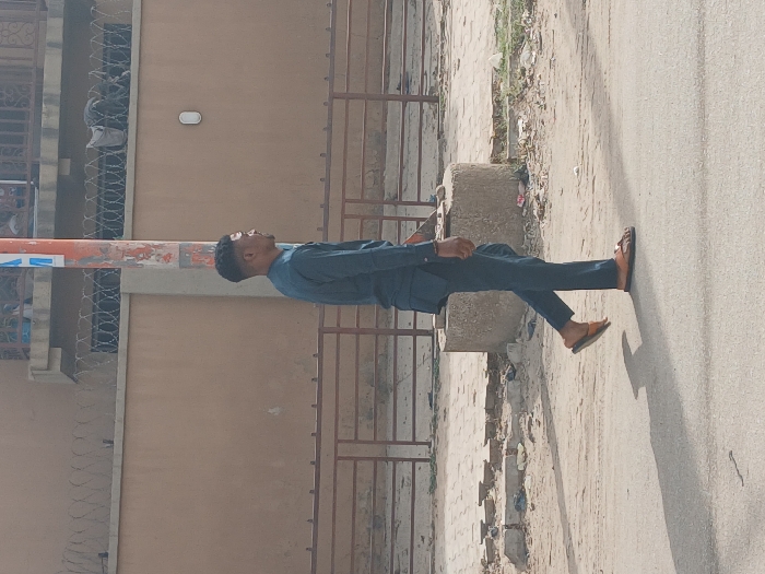 A male individual is depicted in profile, facing right, walking on an asphalt road in Mgbidi, Nigeria. He wears a dark blue long-sleeved tunic and matching trousers, with light brown thong sandals. His left foot is elevated, indicating a striding motion. A distinct shadow is cast on the road to his left.

In the midground, a concrete curb transitions to a dirt and debris-strewn area. A dark brown metal fence, consisting of horizontal bars, runs parallel to the road. Behind this fence, a light brown wall is topped with multiple strands of coiled razor wire. A white oval-shaped light fixture is affixed to the wall. Above the wall, a portion of a building with a dark railing and obscured items is visible.

A weathered orange and white pole with peeling paint stands vertically in the midground, to the right of the subject. Adjacent to the pole, on the ground, is a low, cylindrical concrete structure.