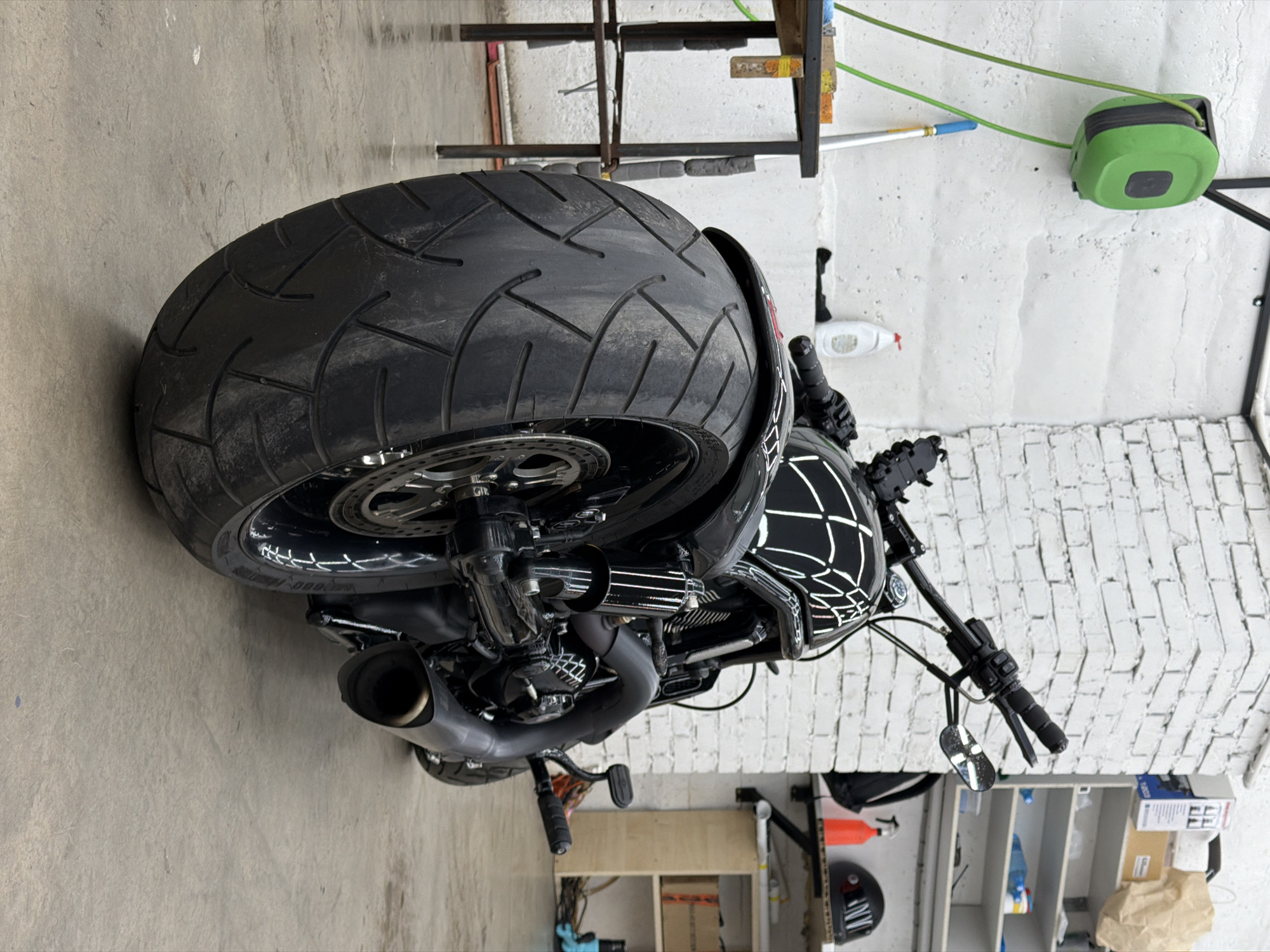 A black custom motorcycle, possibly a cruiser or bobber style, is parked indoors in what appears to be a garage or workshop. The motorcycle is positioned with its rear facing towards the viewer, showcasing its wide rear tire and exhaust system. The fuel tank features a distinctive white web-like graphic pattern against its black surface. The large rear tire has a deep, angular tread pattern, and a metallic disc brake is visible on the rear wheel assembly. The exhaust pipes are black.
The motorcycle is stationary; no activity is taking place. The setting includes a concrete floor and a wall that is partially white painted brick on the lower section and a smoother white surface above. A green hose reel with a green hose is mounted on the wall. Shelving units holding various items, tools, and a white spray bottle are visible in the background. Faint, illegible white text or markings can be seen on the sidewall of the rear tire. The overall scene suggests an indoor storage or maintenance area, consistent with a garage in Krasnoyarsk, Russia, as indicated by the location context.