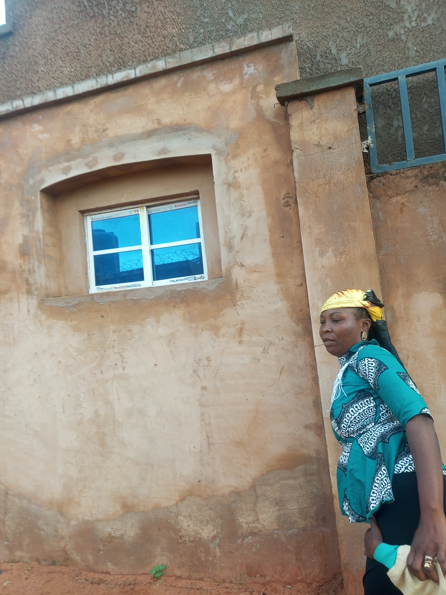 An outdoor, eye-level, medium shot depicts a woman standing to the right of the frame in front of a textured, tan-colored building. The woman, who appears to be in her 40s, is facing to her left and looking away from the camera. She has dark skin and is wearing a yellow headscarf, a green and white patterned top, and black pants. Her left hand is holding a light-colored cloth, and she is wearing rings on her fingers. To the left of the woman is a window with a white frame and blue tinted panes. The window is set into the tan wall, which has some discoloration and texture. The background shows more of the building's exterior and a portion of a metal fence to the right. The ground in the foreground is reddish-brown dirt with some small green plants. The lighting suggests it is daytime.