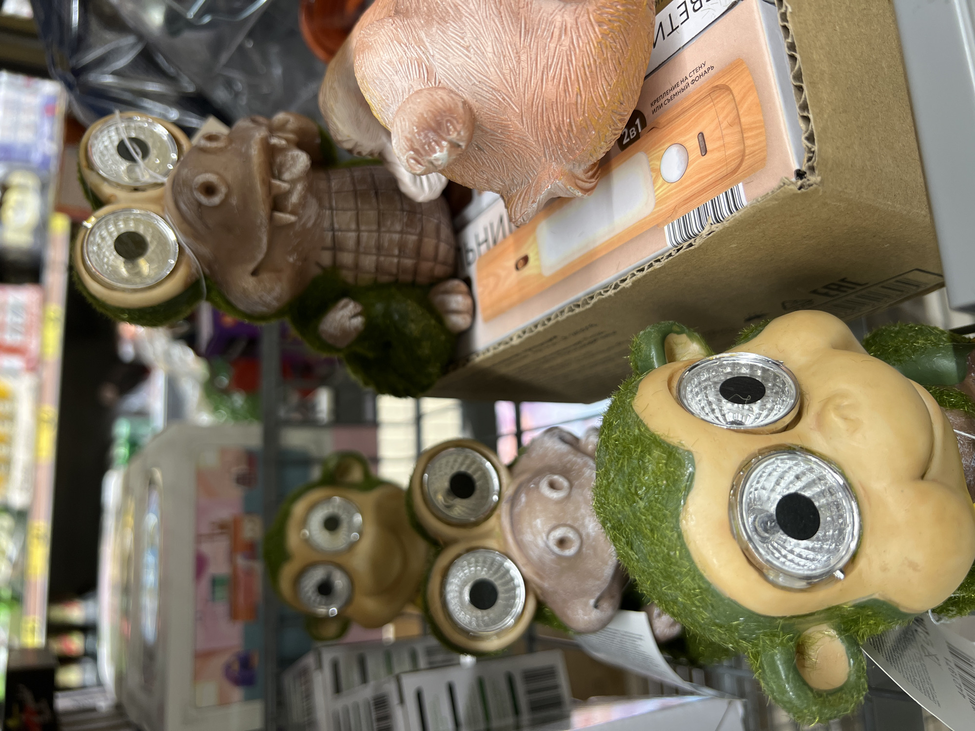 The image is a close-up, medium shot of a retail display featuring novelty garden ornaments. The foreground is dominated by a moss-covered monkey figurine with large, solar-powered eyes, its head tilted slightly to the left. Behind it, stacked vertically, are two more similar monkey figurines, also with solar-powered eyes, and a smaller, light-brown creature with similar eyes. To the right and slightly behind the monkeys is a light-brown alligator figurine with its mouth open, revealing small teeth, and its eyes are also solar-powered. To the far right, partially obscured, is a pink, pig-like figurine. These ornaments are placed in a cardboard box which contains a packaging for a flashlight or similar device with a wood-grain design and some Russian text. The background shows shelves with various retail items, suggesting an indoor store environment, possibly a supermarket or a home improvement store. The lighting is artificial, and the overall impression is one of a commercial display. The location is not identifiable from the image itself.