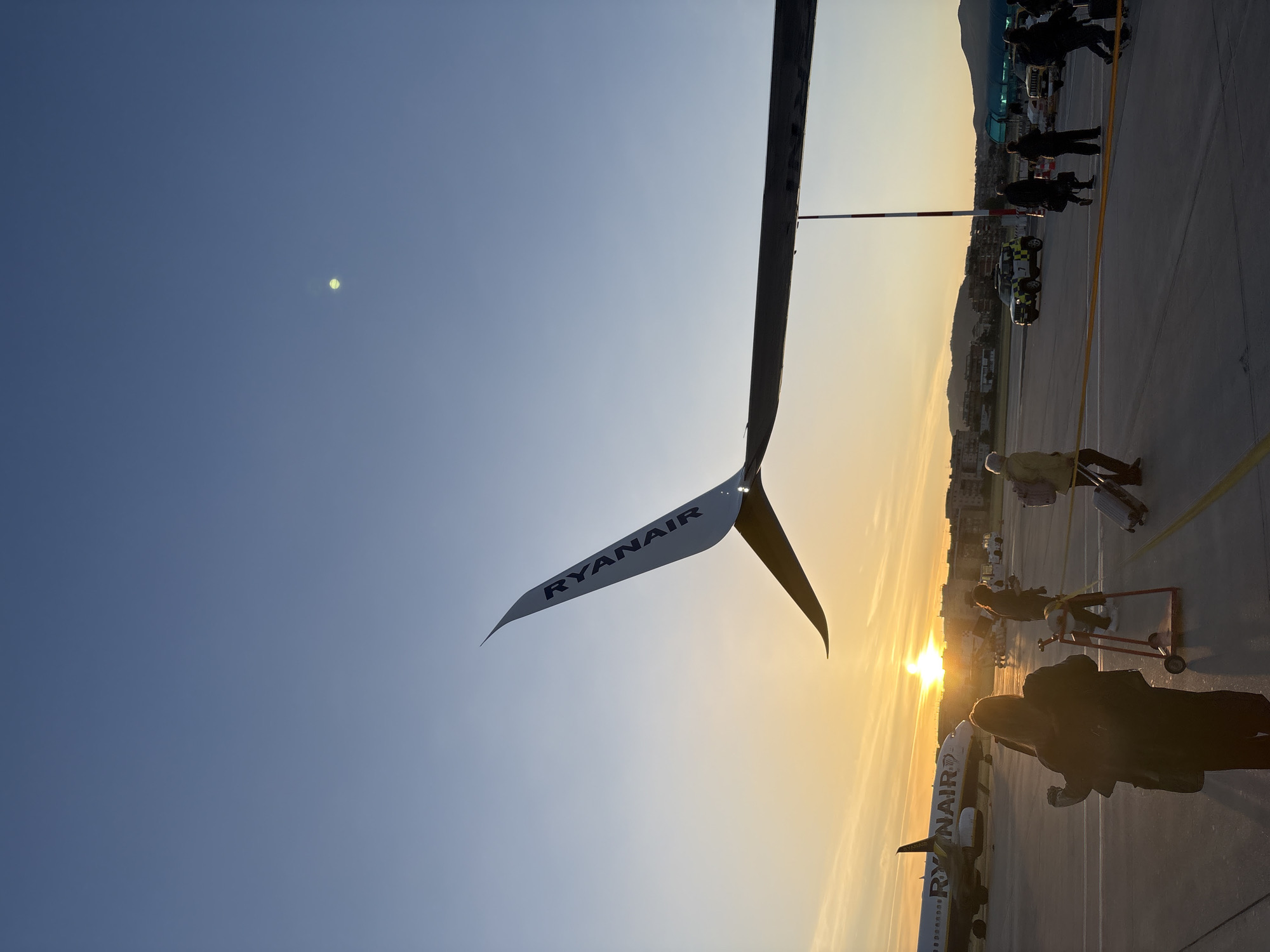 An outdoor scene captures a bustling airport tarmac at either sunrise or sunset, characterized by warm, golden light spreading across the horizon. The image is taken from a low perspective, looking towards the setting/rising sun.
In the upper left quadrant, the distinctive white winglet of a Ryanair aircraft is prominently featured, with the "RYANAIR" logo clearly visible in dark letters on its surface. This wing extends diagonally across the frame, partially obscuring the sky.
On the tarmac below, numerous people are visible, mostly silhouetted against the bright sun. They appear to be passengers, many carrying or pulling luggage on trolleys, suggesting boarding or disembarking activity. Some individuals are wearing high-visibility vests, indicating airport ground staff. Further into the distance, another Ryanair aircraft is partially visible, also bearing the "RYANAIR" livery on its fuselage, and appears to be parked or taxing.
Ground support vehicles, including a small white vehicle with "AIRPORT" text and numbers on its side, are present. A red and white striped pole, possibly an airfield marker, stands upright. Mountains or hills rise in the background, framing the airport buildings visible beyond the tarmac.
The sky transitions from a deep blue overhead to vibrant hues of orange and yellow near the horizon, where the sun shines intensely, casting long shadows. The overall impression is one of active airport operations during a beautiful twilight hour in Rome, Italy.