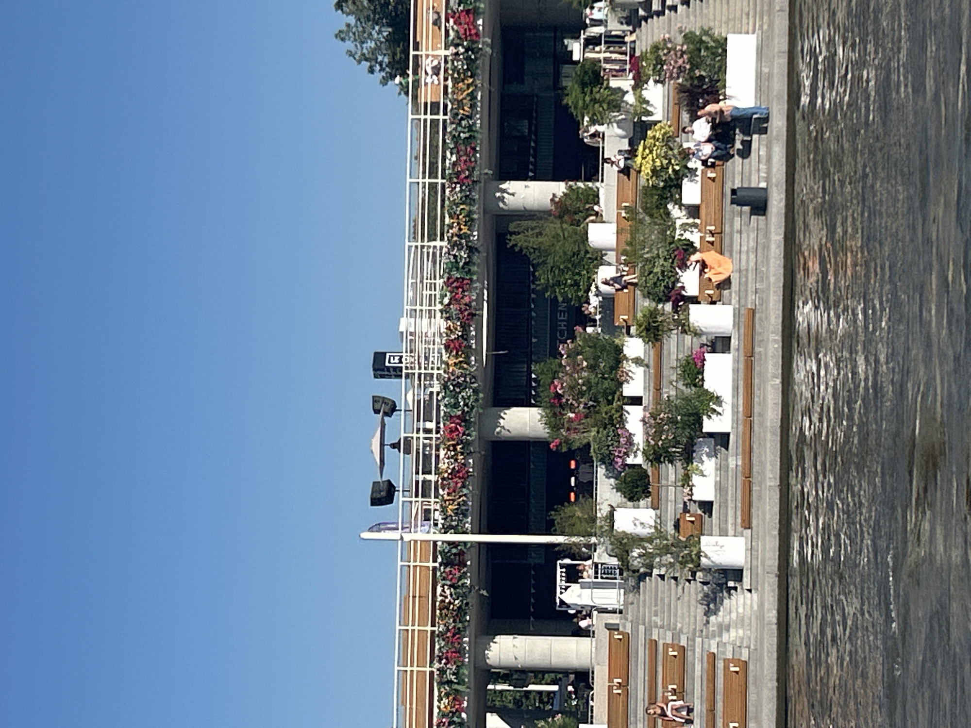 The image shows a multi-level outdoor seating area with a clear blue sky above. The seating is arranged in tiered rows of wooden benches on concrete steps. Numerous white planters filled with various plants and flowers are interspersed throughout the seating area, adding a vibrant splash of color. Some people are visible sitting on the benches and walking along the steps. In the background, there is a modern building with a white railing along its upper level, decorated with hanging floral arrangements. Speakers and an umbrella are visible on the upper level. The scene appears to be a public space, possibly a restaurant or entertainment venue, situated by a body of water, which is visible in the foreground with rippling reflections. The overall atmosphere suggests a pleasant, sunny day at an urban waterfront. This scene is located at the "Luzhniki embankment" on the Moskva River in Moscow, Russia, adjacent to the Luzhniki Stadium complex.