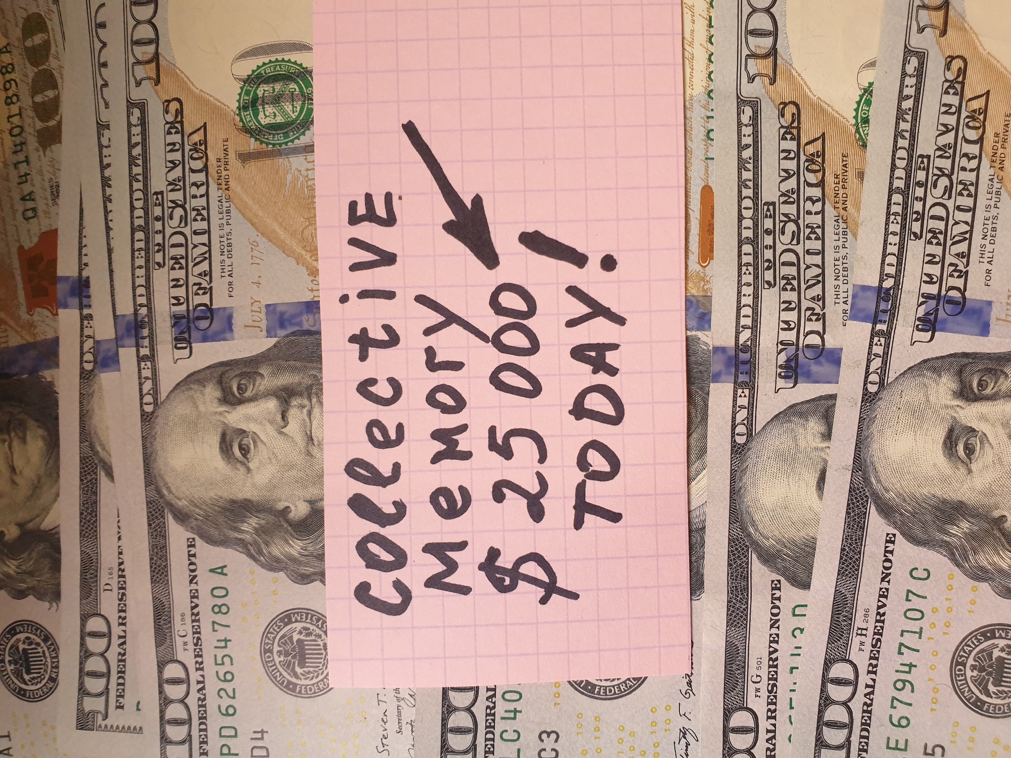 The image shows a pile of several United States one hundred dollar bills. A square piece of pink grid paper is placed over the bills. Written on the paper in black marker is the text "collective memory $25,000 TODAY!" An arrow points downward toward the monetary amount. The scene takes place in an unknown location.