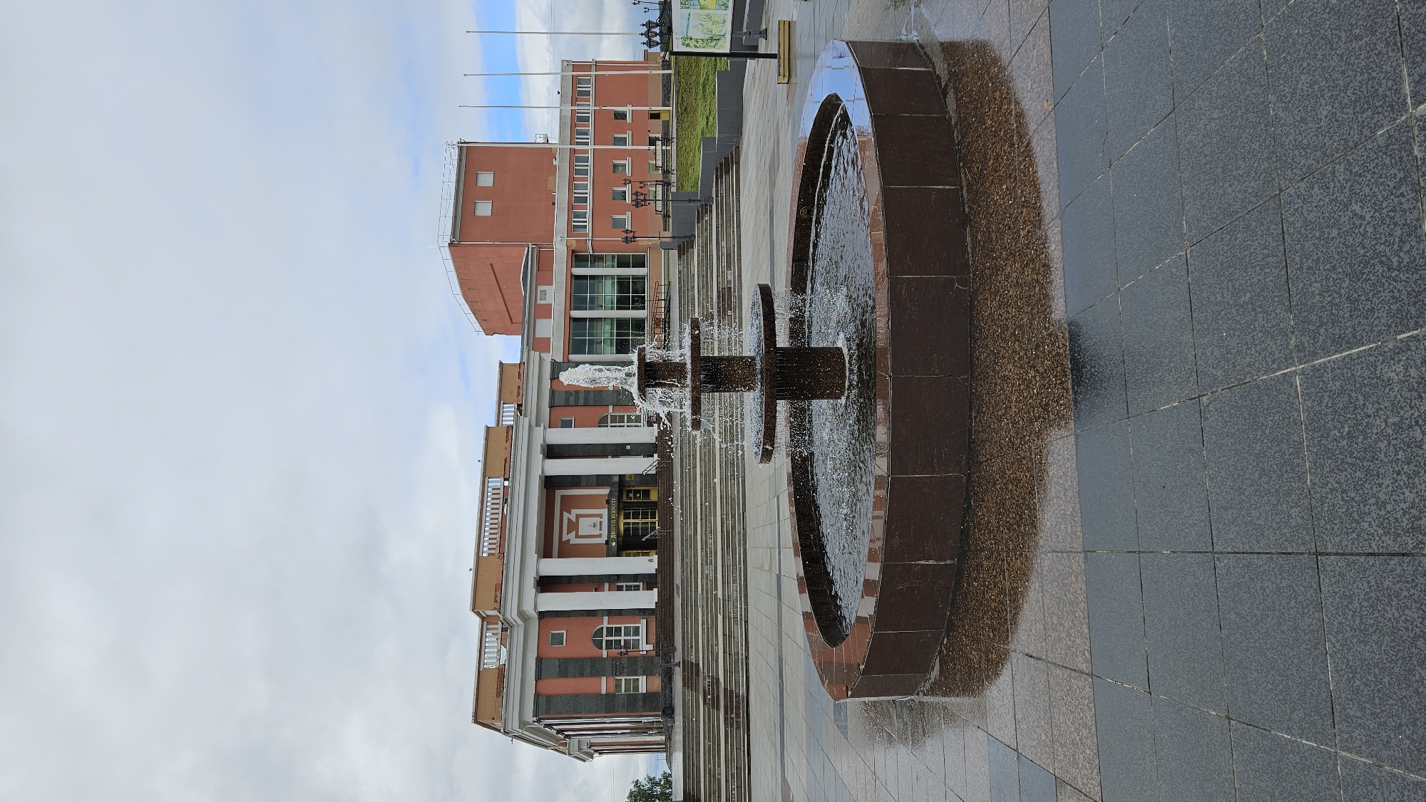 A tiered fountain with water spraying from its top is positioned in the foreground of the image, situated in a paved area with a dark tiled surface. The fountain is surrounded by a circular, dark-rimmed basin. In the background, a large, multi-story building with classical architectural elements is visible. This building features a salmon-pink facade, white columns, a grand staircase leading to its entrance, and a large, modern extension with glass windows. The sky above is overcast with gray clouds. The overall scene appears to be an urban public space.