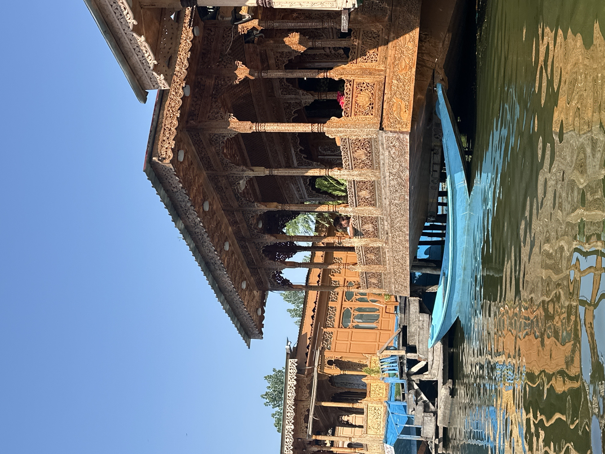 This image depicts a scene on Dal Lake in Srinagar, India, focusing on the intricate wooden architecture of several houseboats. The foreground shows the calm, rippling water reflecting the warm hues of the houseboats and the clear blue sky. A bright blue wooden boat is moored alongside the houseboats.
The houseboats themselves are elaborately decorated with detailed wood carvings. The main houseboat in the right foreground features a covered porch with ornate columns and a railing adorned with floral motifs. The houseboats to the left exhibit similar craftsmanship with detailed wooden facades, wooden decks, and windows. Blue tarps and wooden steps are visible connecting the houseboats and the water. Trees are visible in the background, suggesting a natural setting. The clear sky indicates a sunny day.
