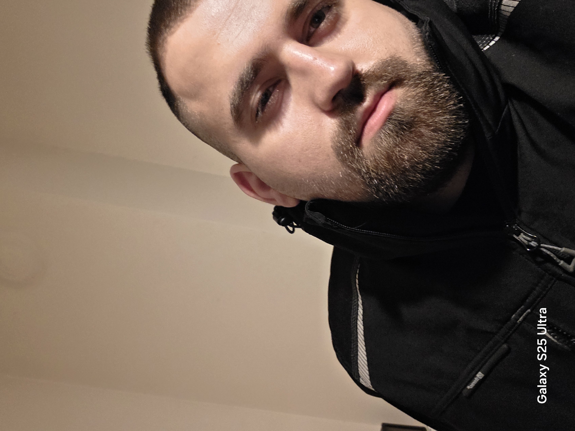 A close-up selfie taken indoors shows a man with a short, dark haircut and a full beard. He is wearing a black jacket. The background is a plain, light-colored wall. The image includes a watermark indicating it was taken with a Galaxy S25 Ultra. There is no information to suggest the picture was taken in Plovdiv, Bulgaria.