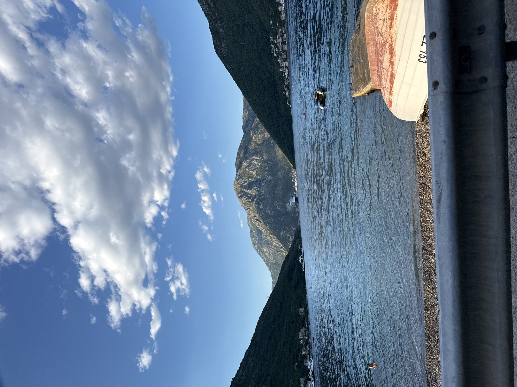 The image depicts a serene coastal scene in Jošice, Montenegro, bathed in the warm light of a late afternoon sun. A tranquil body of water, likely the Bay of Kotor, stretches out towards imposing, verdant mountains that frame the horizon. These mountains, dotted with small villages clinging to their slopes, cast long shadows, adding depth and dimension to the landscape.
In the foreground, a small, pebbled beach meets the calm water. Two inverted boats rest on the shore, one with visible markings that appear to read "CL 153". A concrete jetty extends into the water, showing signs of wear and tear. A person is visible swimming in the distance, a solitary figure enjoying the refreshing embrace of the sea. Further out, a small inflatable dinghy floats on the water, its presence adding to the sense of leisurely activity.
The sky above is a vibrant blue, adorned with a scattering of fluffy white clouds that catch the sunlight, creating a picturesque contrast. The overall atmosphere is one of peace and natural beauty, a typical vista found in this charming Montenegrin locale.