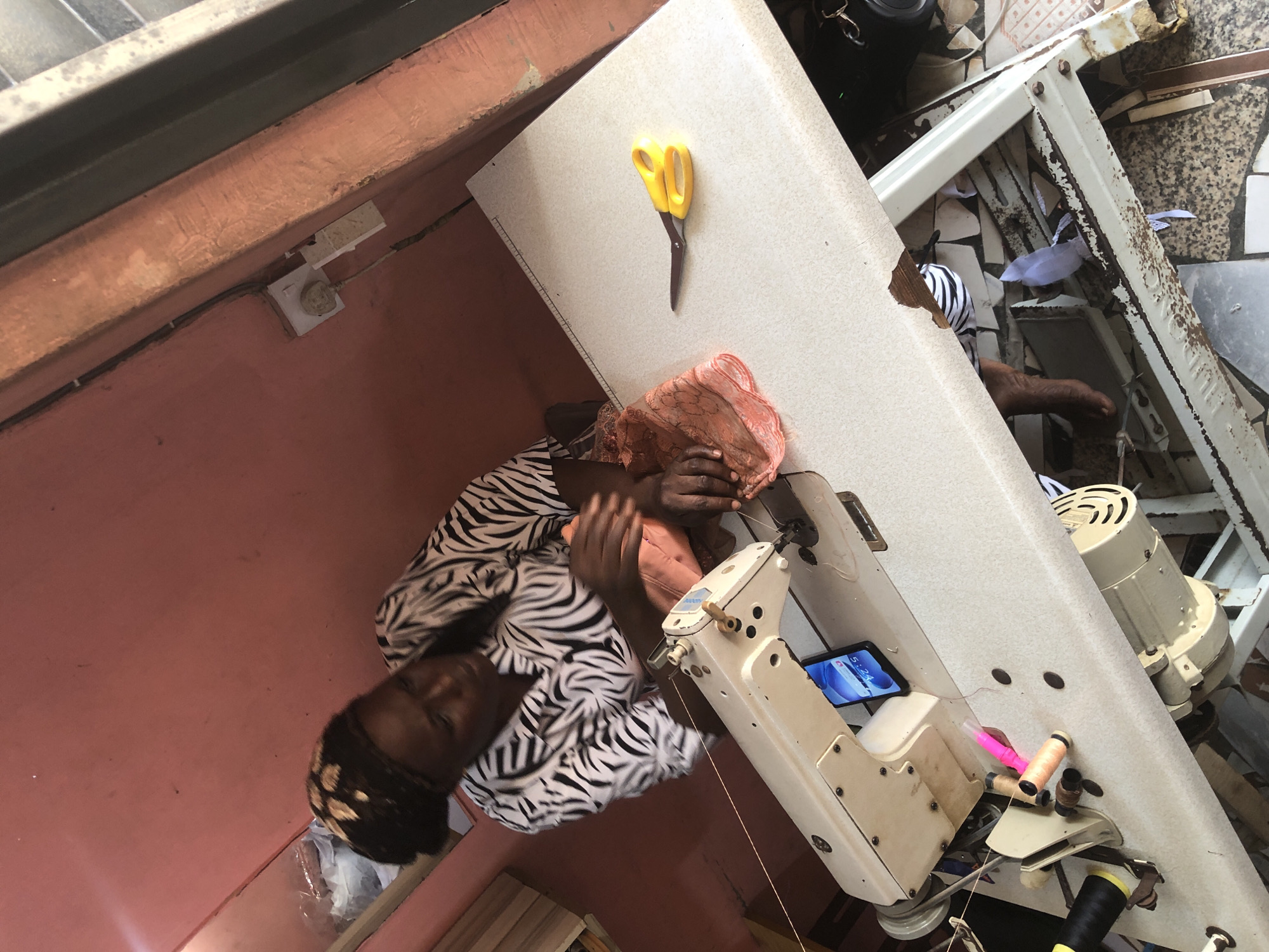 In Ado, Nigeria, a tailor works at her sewing machine. She wears a black and white zebra-print shirt and a headwrap. The sewing machine is white and sits on a beige table. A pair of yellow-handled scissors lies on the table to the right of the machine, and a piece of orange fabric is to the left. A mobile phone is propped up on the sewing machine, displaying the time "5:24." Several spools of thread, including a pink one and an orange one, are visible on the sewing machine. The background is a reddish-pink wall. The scene is lit by overhead lighting.