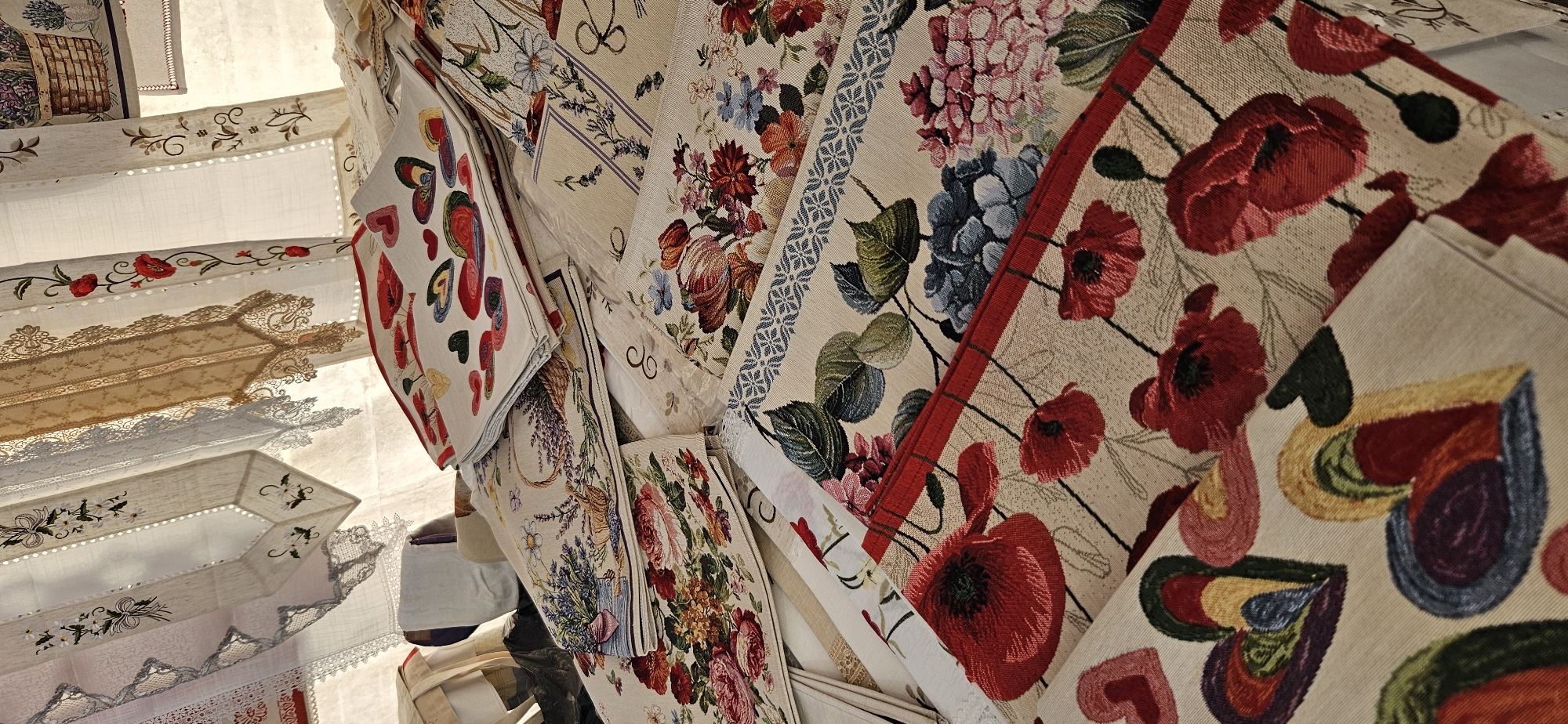 The image displays a collection of embroidered textiles, likely at a market stall in Hévíz, Hungary. The textiles are folded and stacked, featuring various floral patterns, including poppies, roses, and lavender in baskets, as well as heart motifs. Some of the textiles also have decorative lace trim. In the background, sheer fabric panels with embroidered details are visible, suggesting they might be part of a stall canopy or display. There are no people or discernible actions depicted. The lighting is diffused, creating a soft atmosphere.