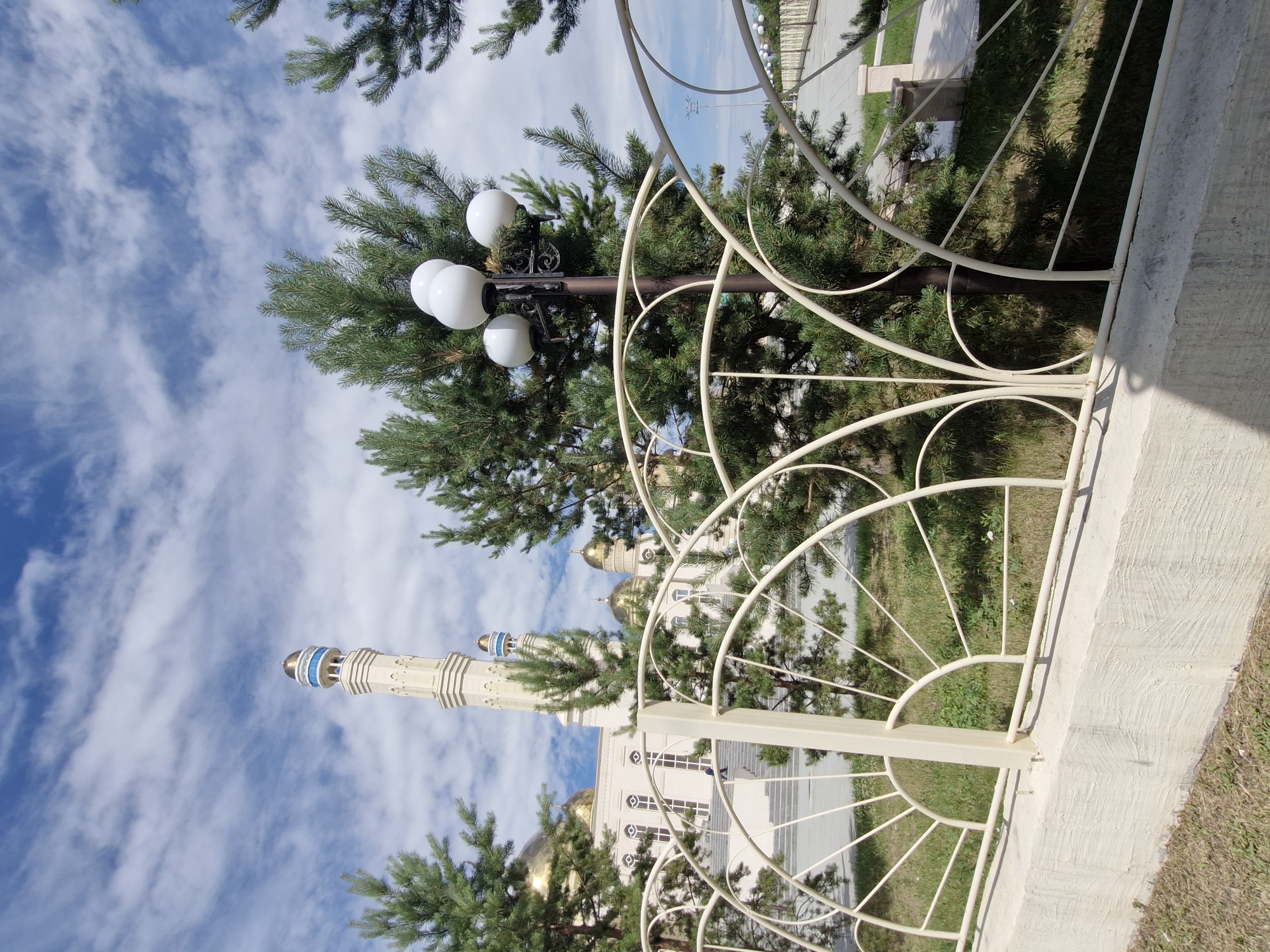 The image depicts a bright, daytime scene with a clear blue sky featuring scattered white clouds. In the foreground, a cream-colored, decorative metal fence runs horizontally across the frame, partially obscuring the view beyond. This fence is mounted on a textured concrete base. Behind the fence, lush green pine trees are visible, their branches reaching upwards. A multi-globe lamppost stands among the trees, its white globes brightly lit.
Further back, a mosque with a tall, cream-colored minaret and golden domes is the dominant structure. The mosque features a light-colored facade and arched windows. A person, appearing as a small dark silhouette, is seen walking up a wide staircase leading to the mosque's entrance. The area around the mosque appears to be a well-maintained landscape with grass and pathways.
Based on available information, this mosque is identified as the Nur-Astana Mosque, a prominent landmark in Kokshetau, Kazakhstan. The image captures a portion of the mosque complex and its surrounding grounds, suggesting a public space open for visitors. The overall impression is one of peacefulness and architectural grandeur set against a natural backdrop.