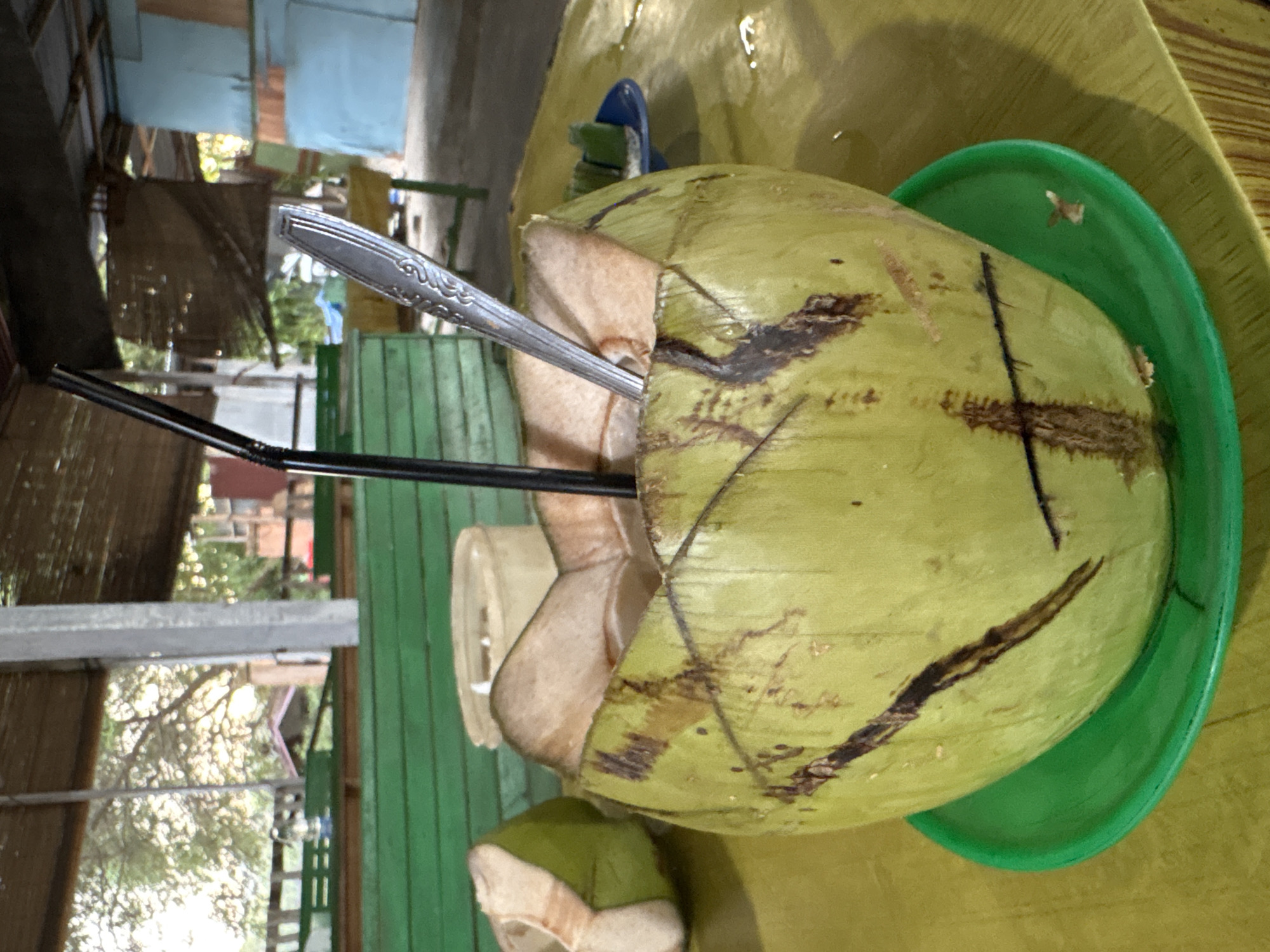 A close-up, eye-level shot shows a fresh green coconut, hollowed out and filled with water, resting on a green saucer on a yellow table. A black drinking straw and a silver spoon are inserted into the coconut. In the background, out of focus, is a partially opened coconut. Further back, a green wooden bench is visible, with trees and a building in the distance. The scene is likely outdoors, possibly at a roadside stall or small cafe in Lhokseumawe, Indonesia, given the tropical foliage and simple architecture.