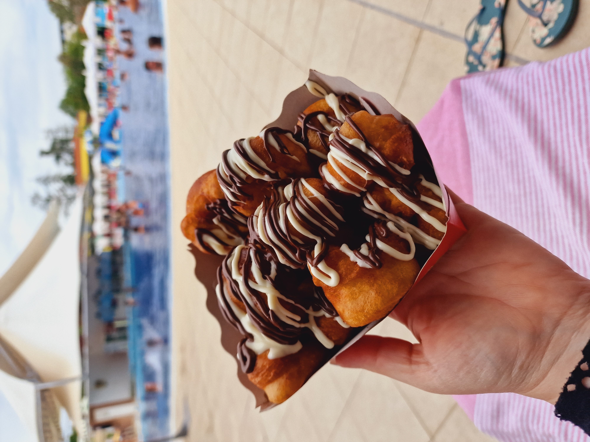 A hand holds a red cardboard tray filled with golden-brown fried dough pastries drizzled with dark chocolate and white icing. The pastries are stacked in the tray and appear to be mini donuts or similar fried dough balls. The background is out of focus and shows a swimming pool area with people swimming and relaxing. A poolside bar with white tents is visible in the distance. The ground is paved with light-colored tiles. A pink and white striped towel and a pair of floral flip-flops are visible in the foreground to the right of the hand. This scene appears to be at a resort or water park, and while the location is mentioned as Kazanlak, Bulgaria, there are no specific visual cues in the image to confirm this.