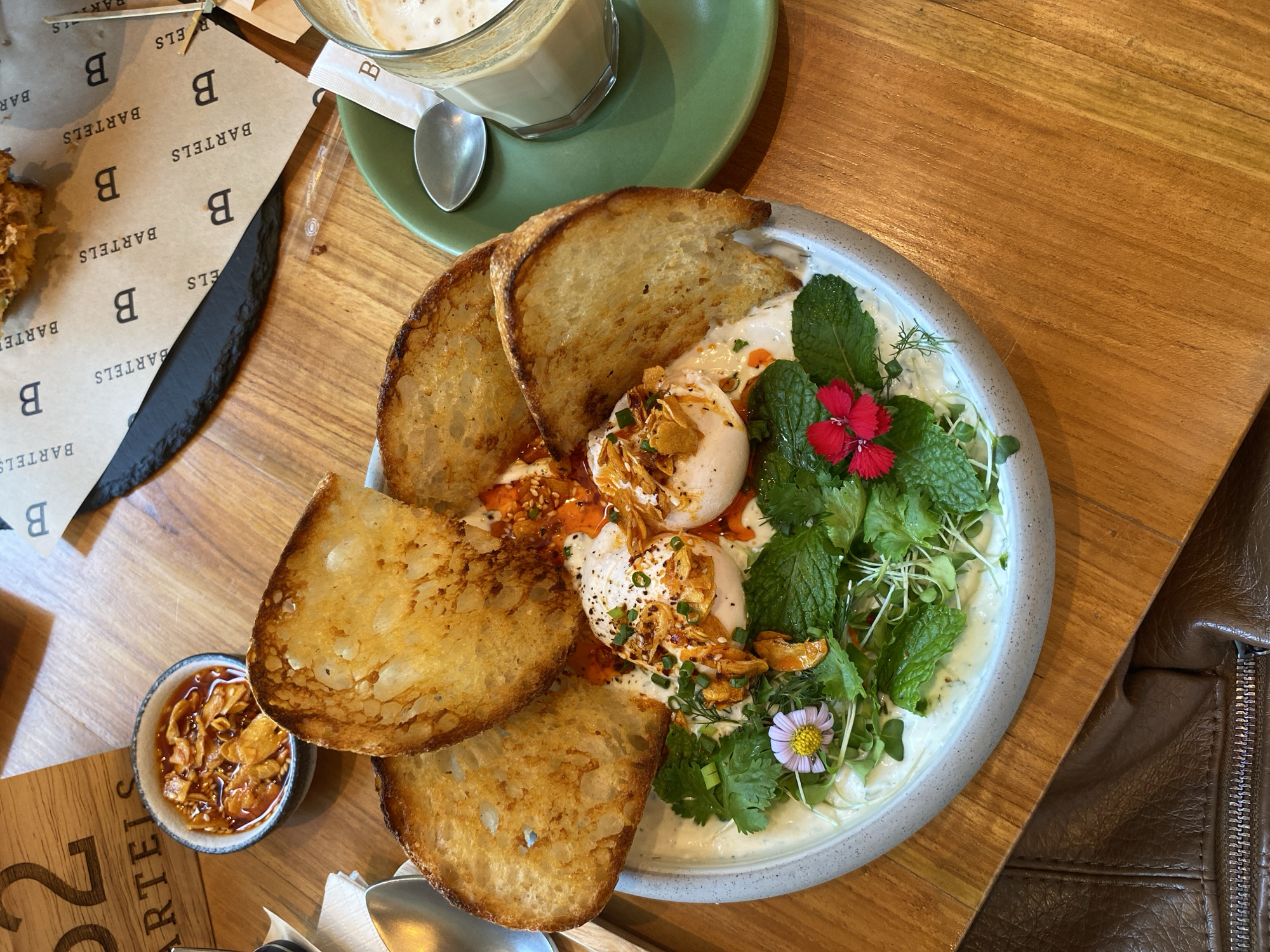 Here is a description of the image:
**Scene:** A high-angle shot captures a wooden table setting with several items related to a meal.
**Food:** The main feature is a plate holding poached eggs, herbs (including mint), what looks like a yogurt or labneh base, and several slices of toasted bread arranged around the plate. A small bowl containing an unidentified garnish is placed near the plate.
**Drink:** A drink, possibly a latte or similar beverage, is in a glass on a saucer.
**Branding:** Papers with the name "Bartels" are scattered on the table. There's also a wooden block with "Bartels" and the number "22" visible.
**Context:** Given the branding elements, it seems the food is being served at a restaurant or café called Bartels.
**Environment:** A brown jacket is partially visible in the bottom-right corner of the frame.
There is nothing in the image that helps identify the location as Bangkok, Thailand.