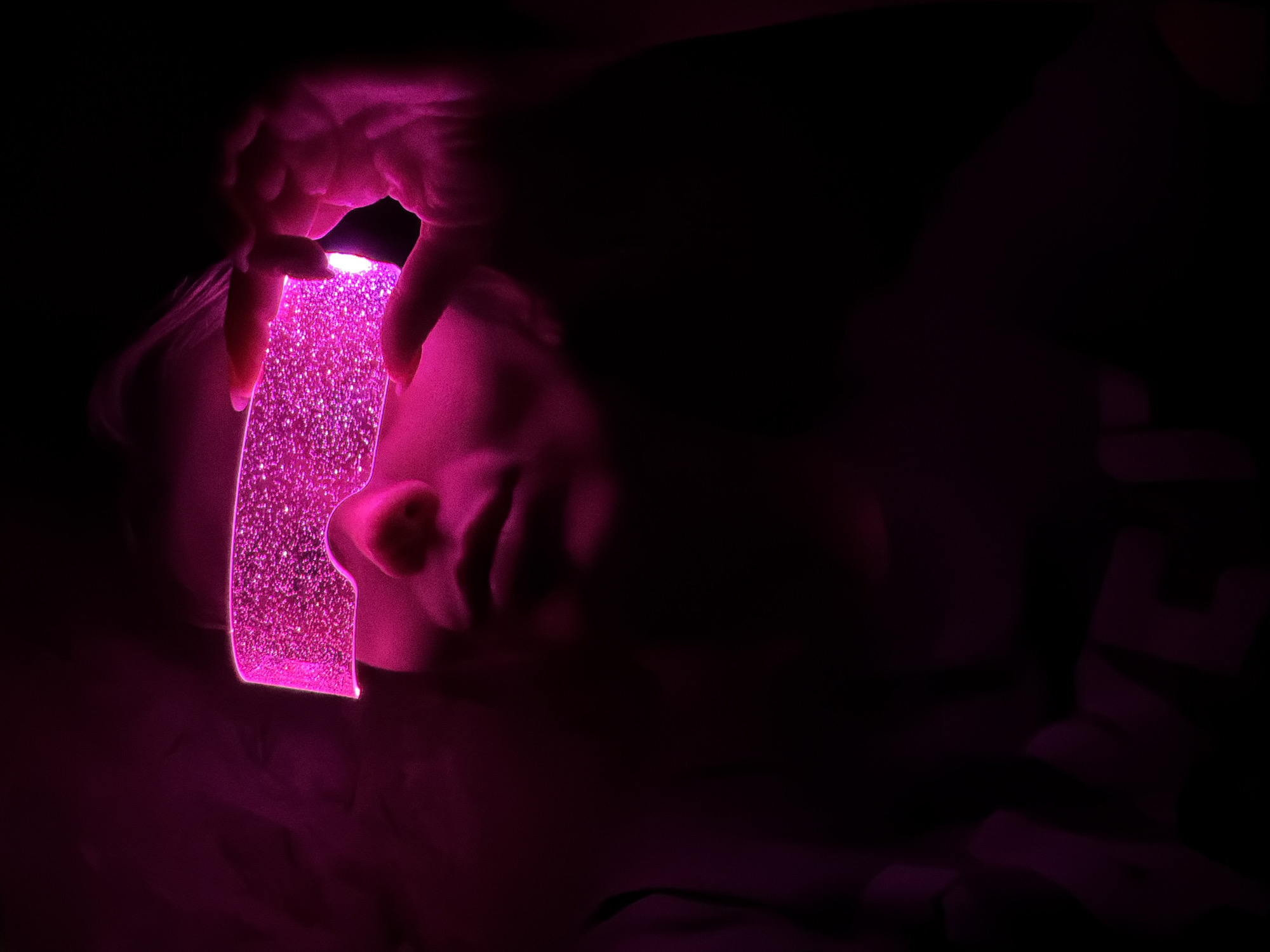 The image depicts a person holding a brightly glowing, pink rectangular object in a very dark indoor setting. The ambient light is almost entirely absent, making the fuchsia glow of the object the sole significant illumination source.
A person, likely a child or young adult, is visible in the left-center of the frame, lying down. Their head and hands are lit by the vibrant pink light. Their hands are holding the luminous object directly above them. The object itself is a translucent, rectangular slab with rounded edges, appearing to be filled with numerous tiny, sparkling particles or glitter suspended in a liquid, creating a dynamic, bubbly effect when lit.
The background is plunged into deep shadows, showing only faint outlines and hints of dark purple where the pink light softly diffuses. This strong contrast emphasizes the glowing object and the illuminated parts of the person. The scene suggests a calm and intimate moment, possibly at night, with the person interacting with a light-up toy or decorative lamp. There is no visible text in the image. The provided location context for the scene is Chaikovsky, Russia.