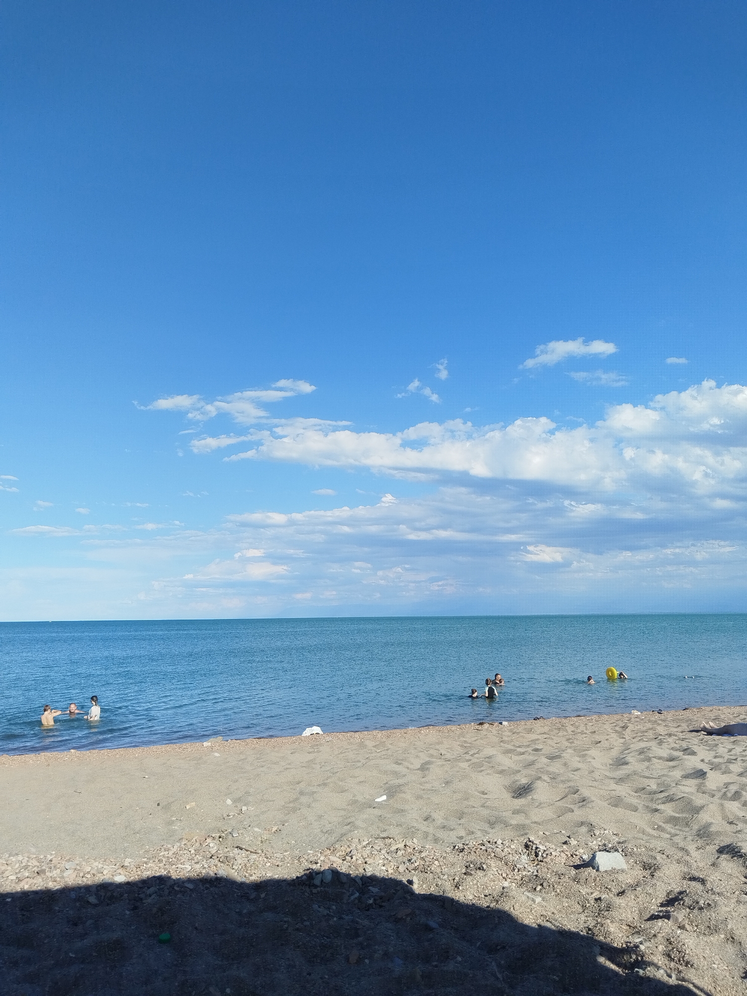 The image depicts a serene beach scene with a vast expanse of calm, blue water meeting a sandy shore under a clear blue sky dotted with scattered white clouds. Several individuals are visible in the water, engaging in recreational activities. On the left side of the water, three people are present, appearing to swim or cool off. Further to the right, a yellow inflatable ring is visible, with a person nearby. The beach itself is composed of sand and small pebbles, with a shadow cast from an unseen object in the foreground, obscuring a portion of the sand. The overall impression is one of leisure and relaxation in a natural setting, likely a popular spot for locals and visitors in the city of Qonaev, Kazakhstan, known for its proximity to the Kapchagay Reservoir which offers similar beach environments.