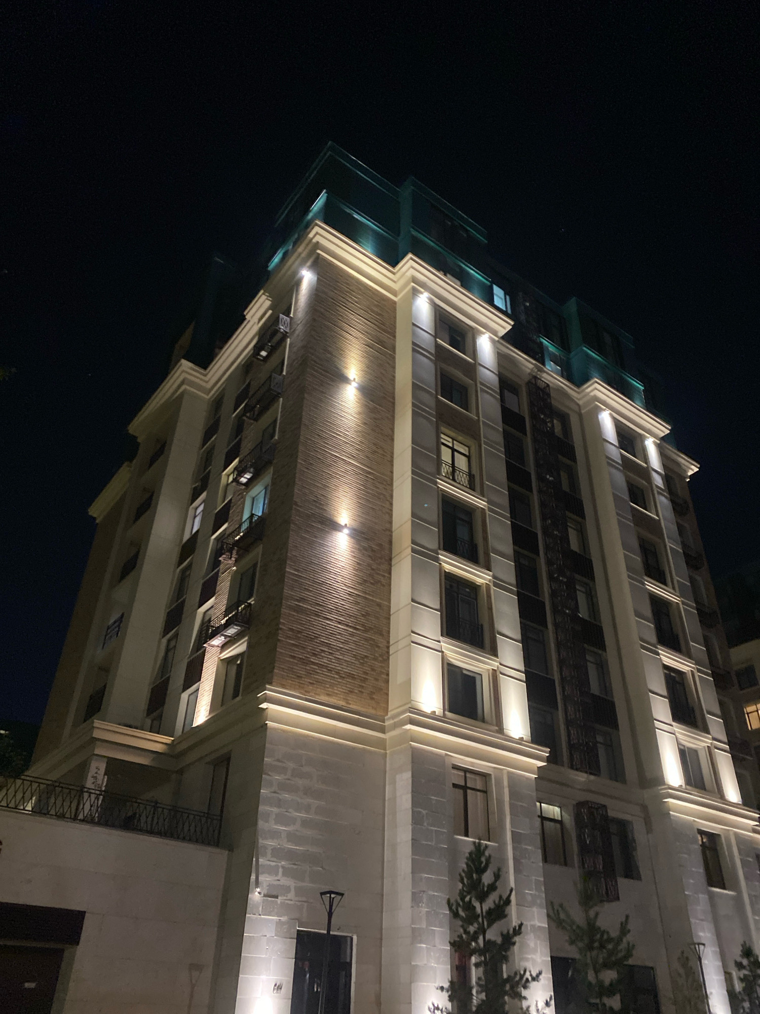 The image shows a multi-story building at night, illuminated by exterior lights. The building has a modern design with a brick facade on one side and a lighter stone-like material on the other. It features multiple windows, balconies with decorative railings, and a green roof. The sky is dark, indicating nighttime. The building appears to be located in a city, with other structures possibly visible in the background, though not clearly identifiable. The image captures the architectural details and lighting of the building.